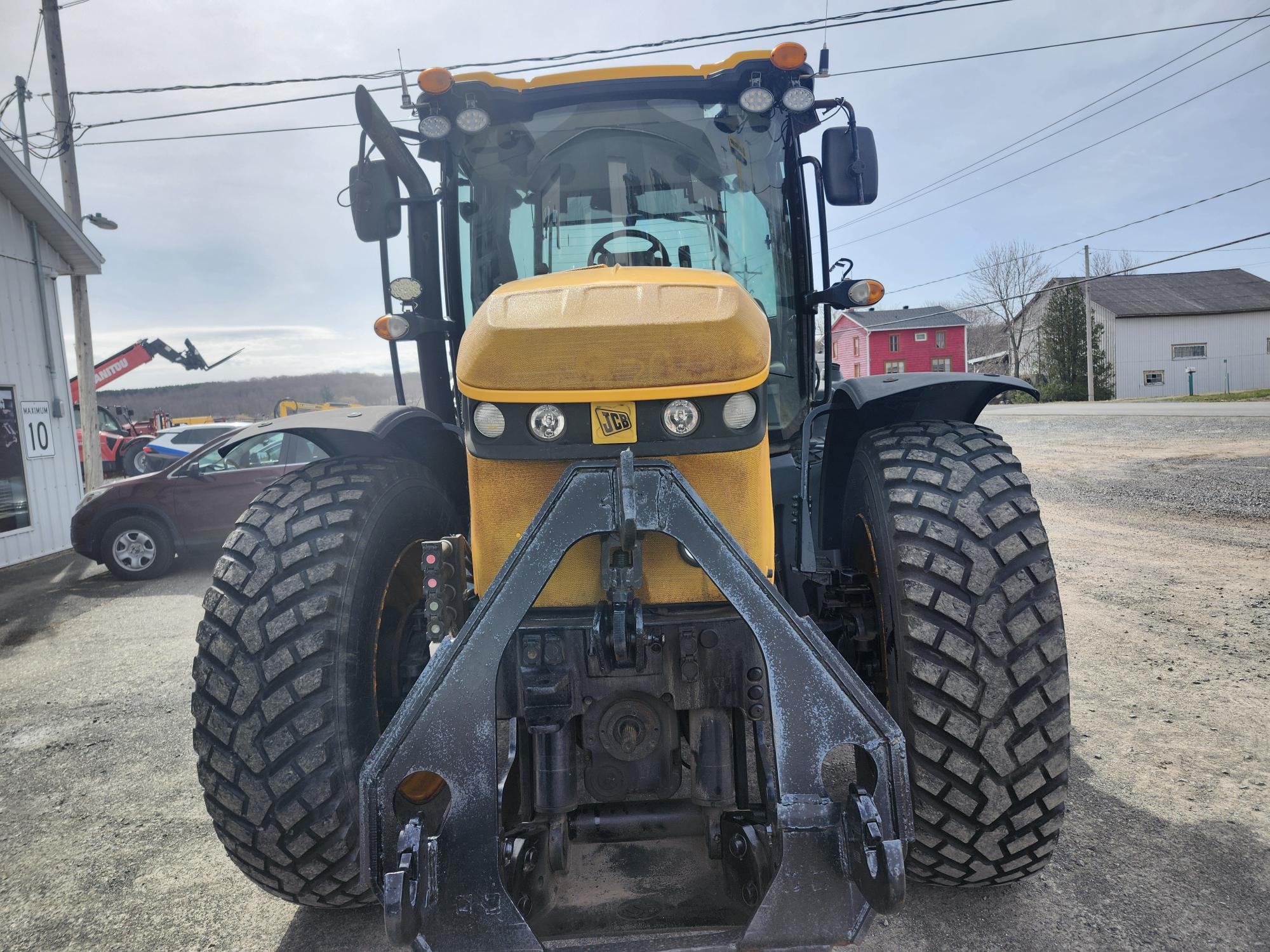 Un gros tracteur jaune vu de face, avec des pneus épais, des phares et un accessoire hydraulique à l'avant. Il est garé sur un terrain pavé avec des bâtiments, des voitures et des lignes électriques visibles en arrière-plan.