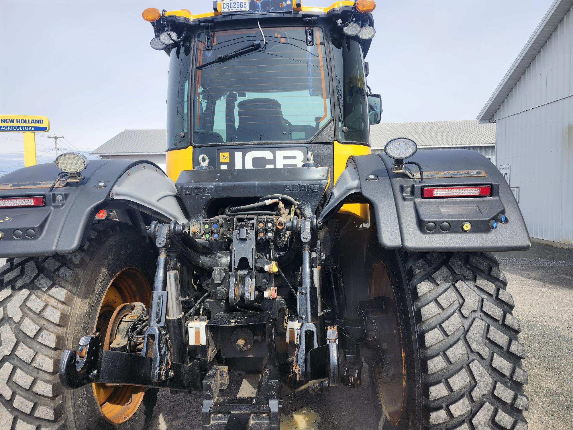 Vue arrière d'un gros tracteur JCB avec des pneus robustes, des composants hydrauliques et un attelage, garé sur du gravier près d'un bâtiment blanc et d'un panneau indiquant « New Holland Agriculture ».