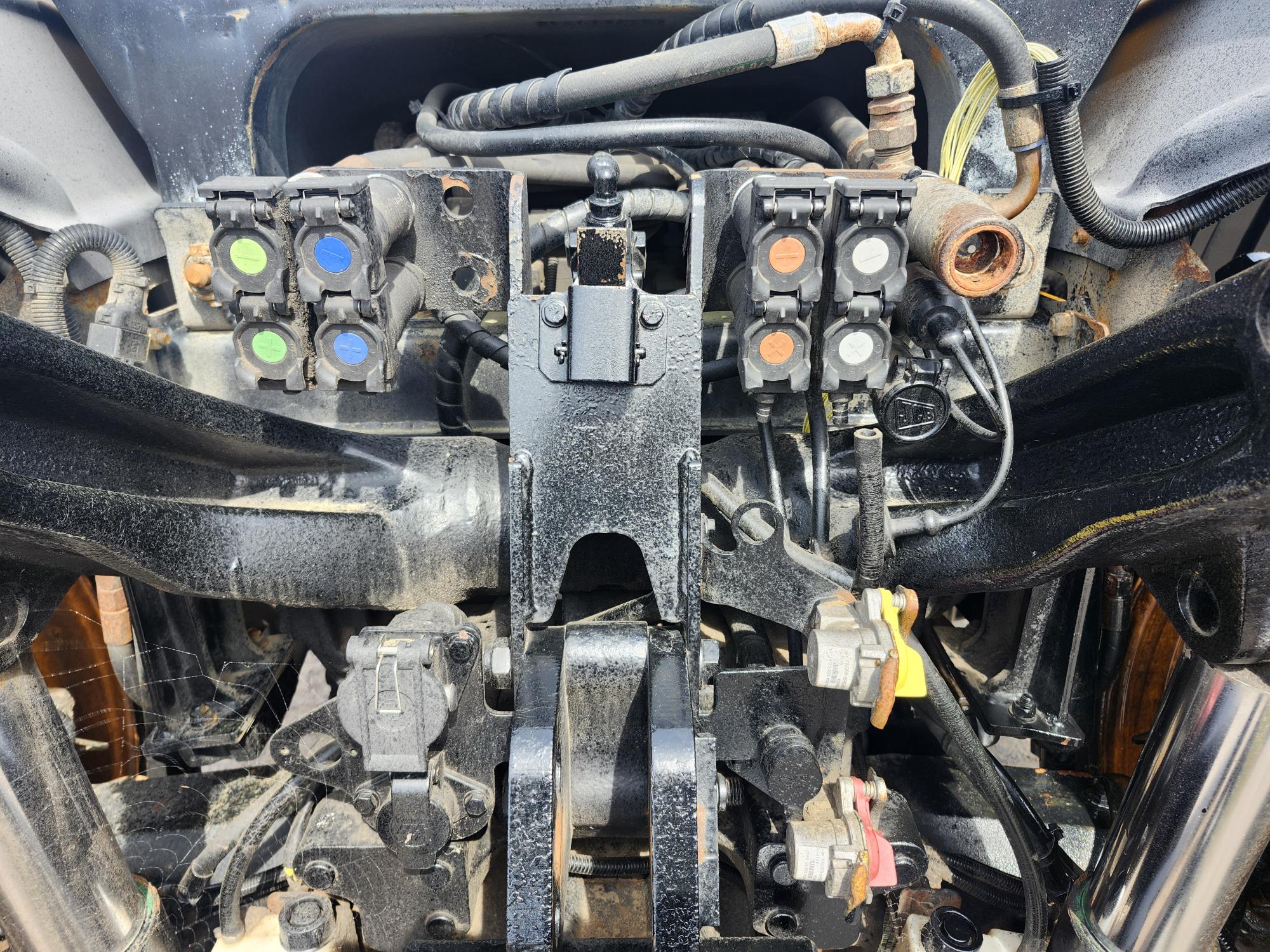 Vue rapprochée des connexions et des raccords hydrauliques arrière d'un tracteur, avec des capuchons colorés pour l'identification, divers composants métalliques, des tuyaux et de la saleté et de la rouille visibles.