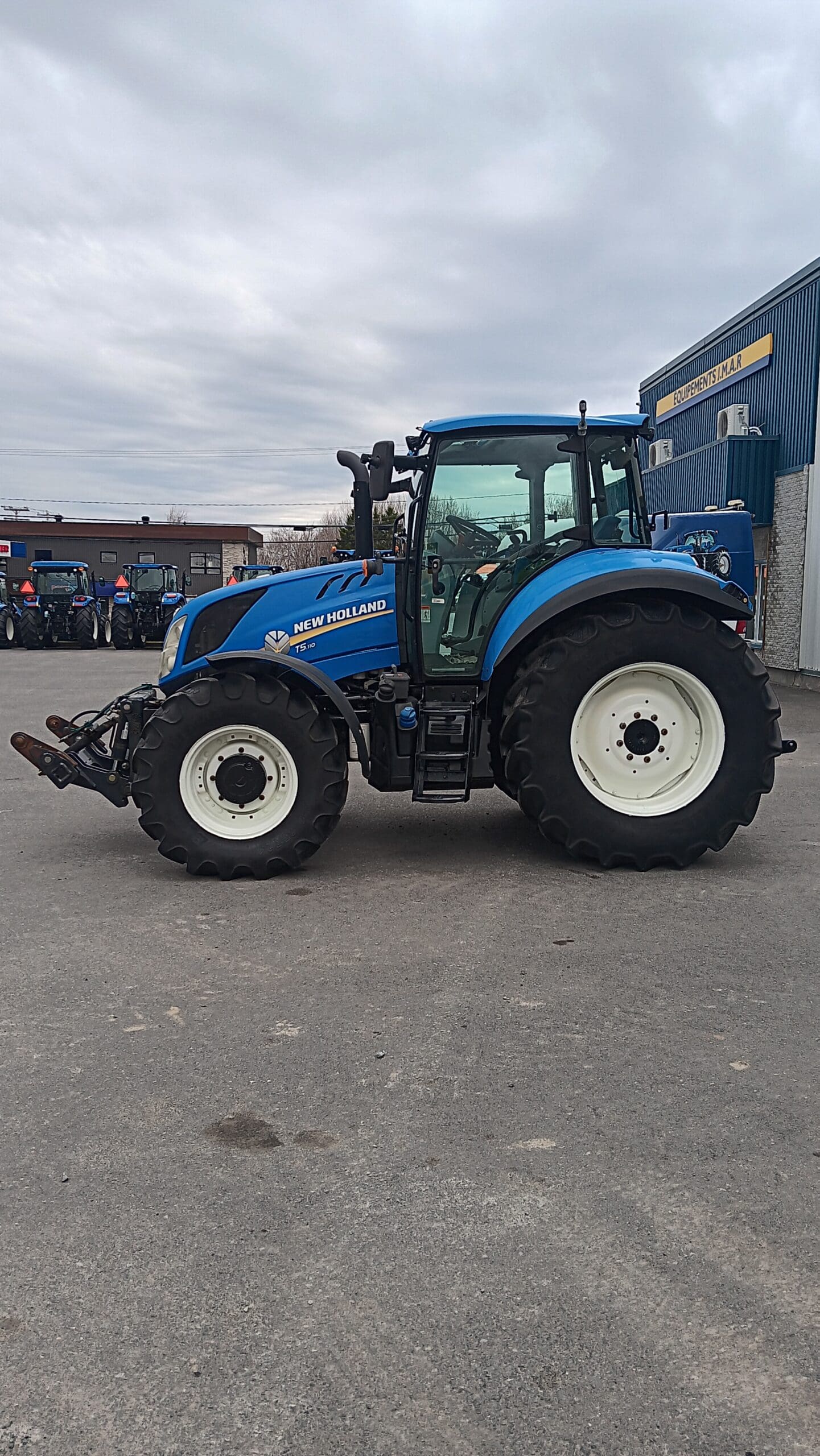 Un tracteur New Holland bleu est garé sur un terrain pavé devant un bâtiment orné d'une pancarte. Le ciel est nuageux et plusieurs autres véhicules sont visibles en arrière-plan.