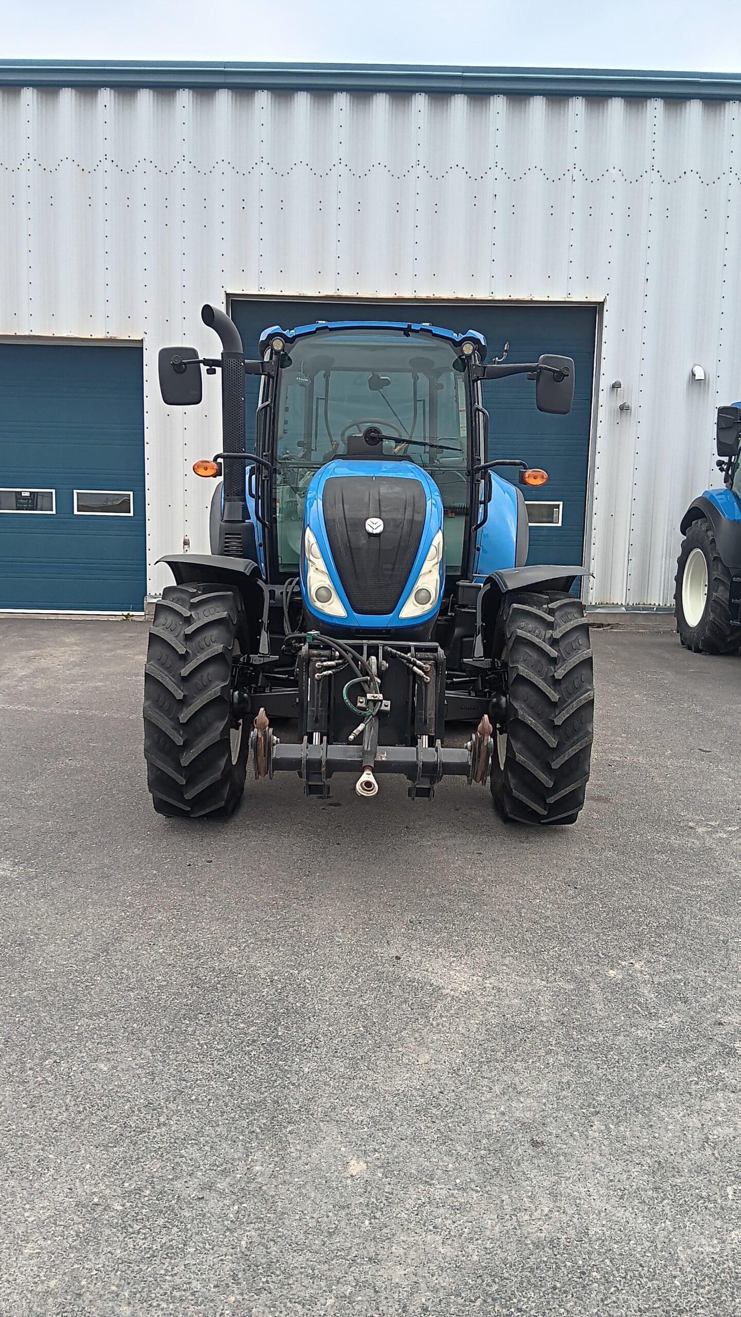 Un tracteur bleu avec de gros pneus noirs est garé sur une surface asphaltée devant un bâtiment en tôle ondulée avec des portes de garage bleues.