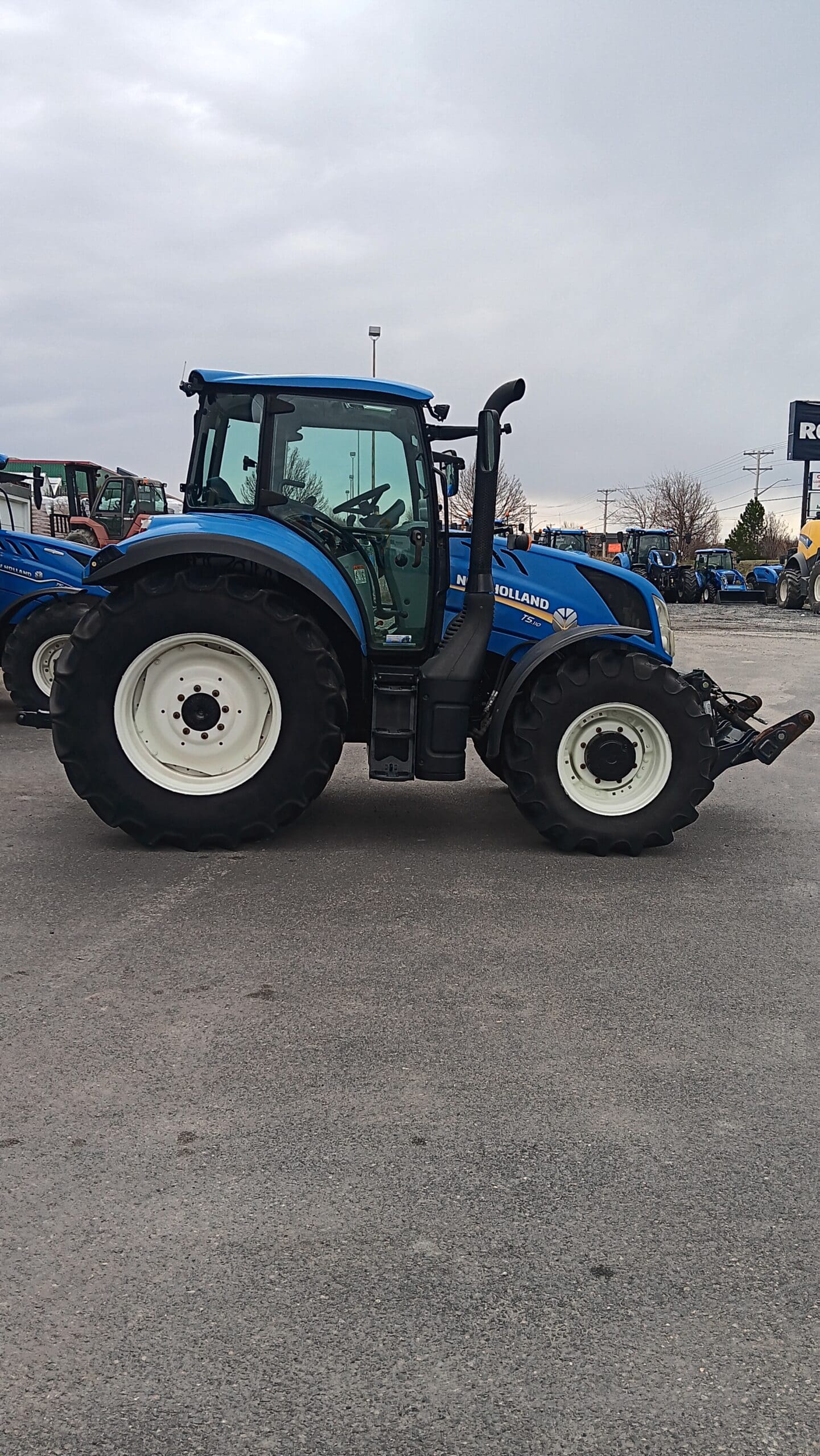 Un tracteur New Holland bleu est garé sur une surface pavée sous un ciel nuageux, avec d'autres tracteurs visibles en arrière-plan.