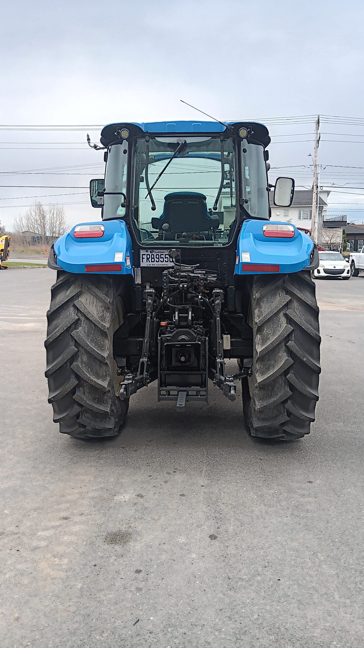 Un tracteur bleu est garé sur une surface pavée, vu de l'arrière. Il est équipé de gros pneus robustes et de machines visibles en dessous. Des voitures, des lignes électriques et des bâtiments se trouvent en arrière-plan.
