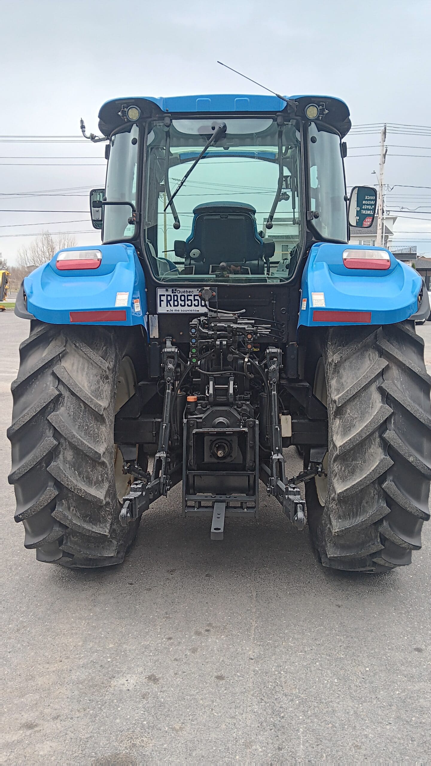 Un tracteur bleu vu de l'arrière, montrant de gros pneus, une plaque d'immatriculation indiquant « FRB955 » et des composants de machines visibles en dessous, garé sur une surface pavée.