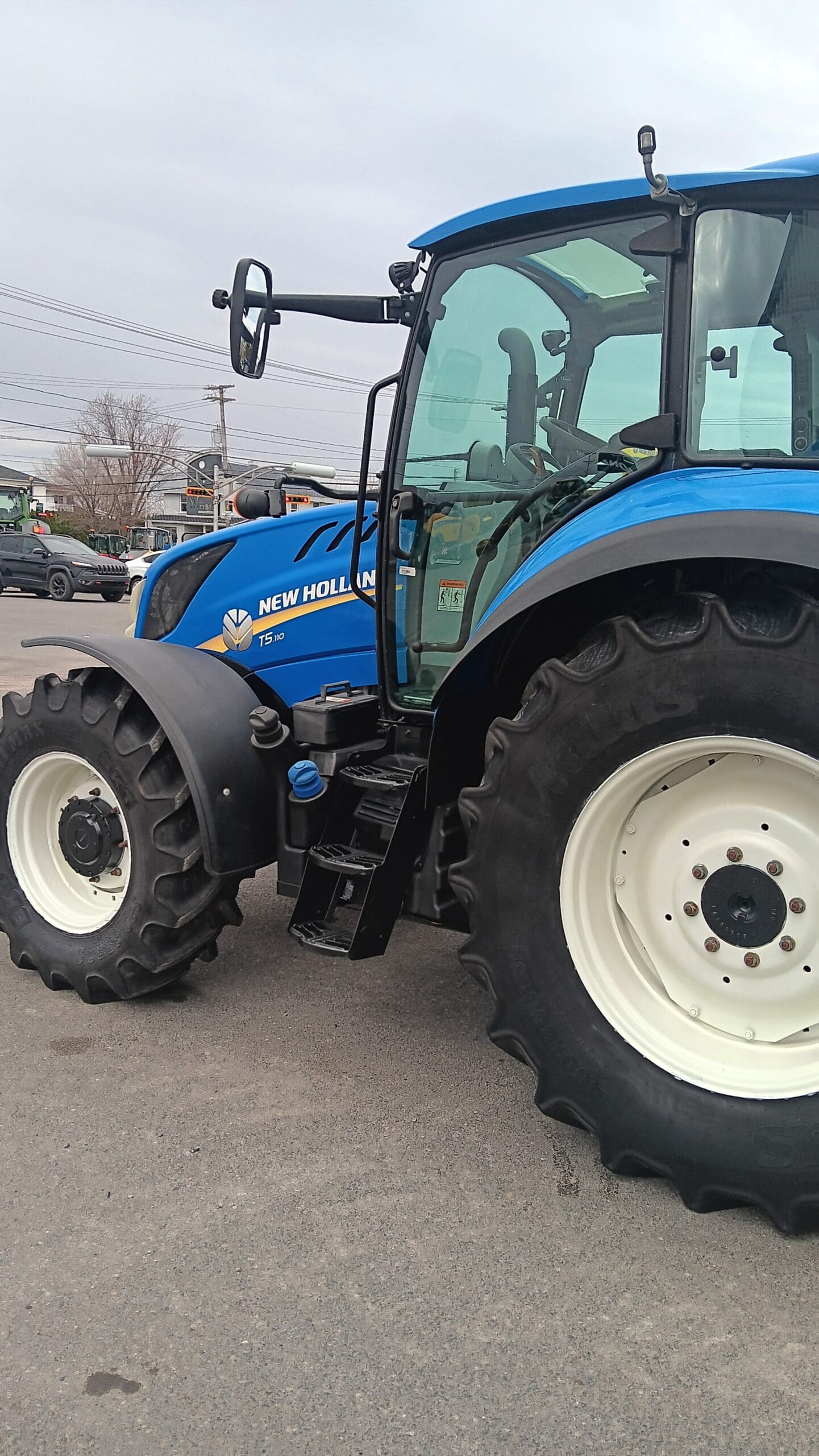 Un tracteur New Holland TS bleu est garé sur une surface pavée sous un ciel nuageux, avec de gros pneus arrière et de petits pneus avant visibles. La porte du tracteur est ouverte, laissant apparaître les marches et une partie de l'intérieur.