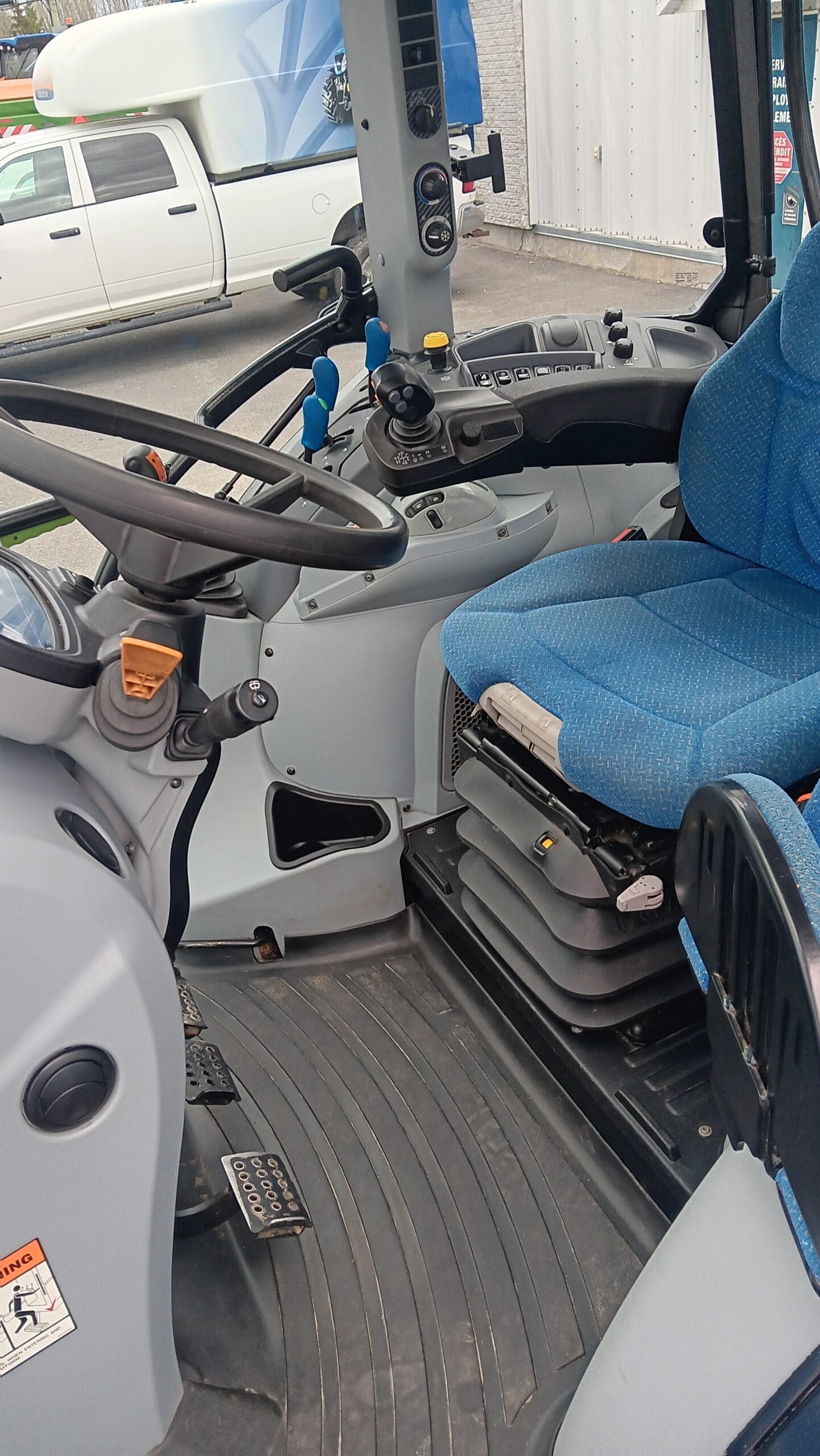 Vue intérieure d'une cabine de tracteur moderne, avec un siège conducteur bleu, un volant, des pédales, des leviers de commande, divers boutons et un tapis de sol gris épuré. Une partie d'un camion blanc est visible par la vitre de la cabine.