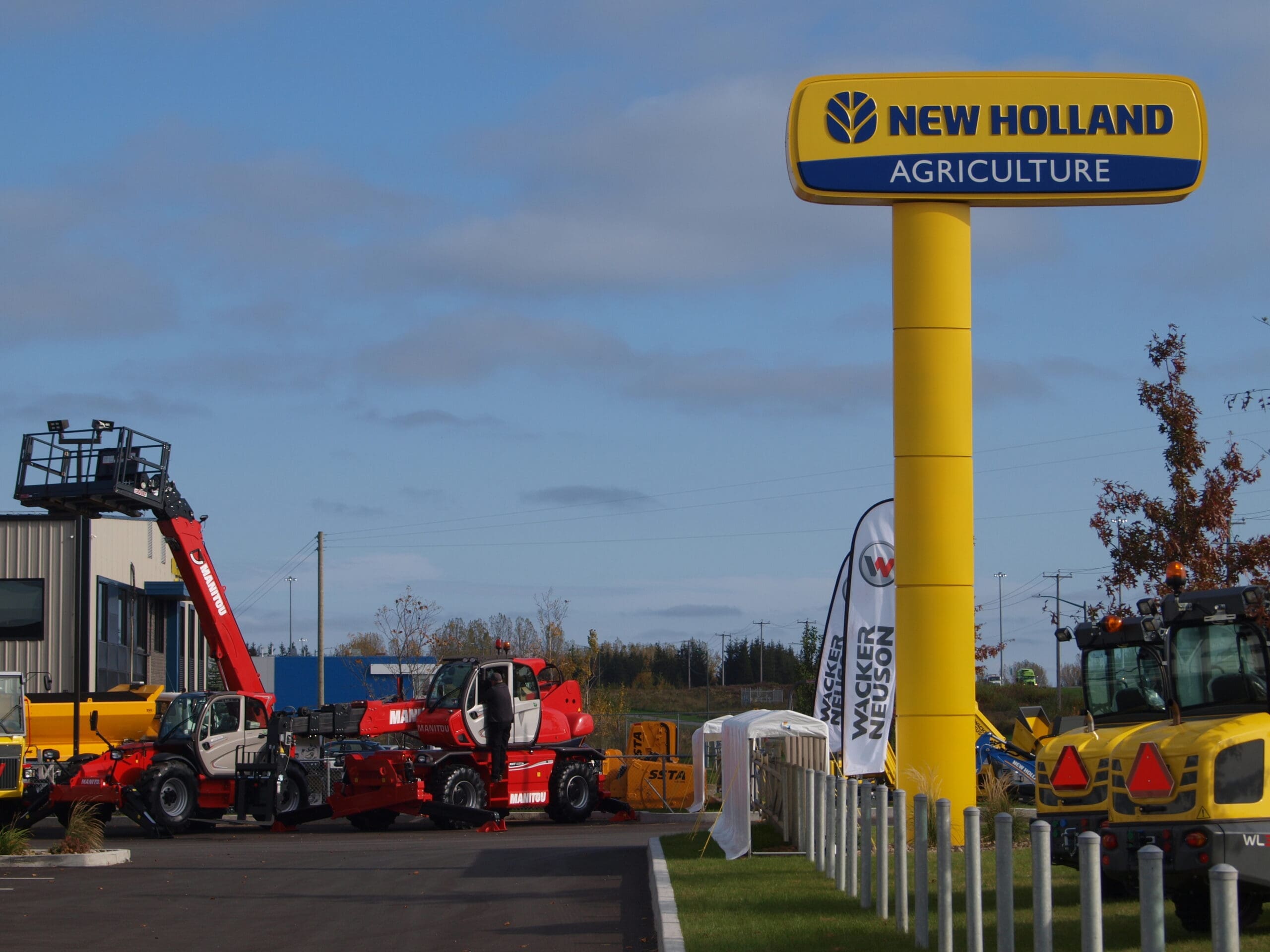 Un concessionnaire New Holland Agriculture exposant divers équipements agricoles, notamment des machines rouges et jaunes, des chariots élévateurs et des tracteurs. Le concessionnaire est identifiable par un grand panneau jaune portant le nom de la marque. Le ciel est partiellement nuageux.