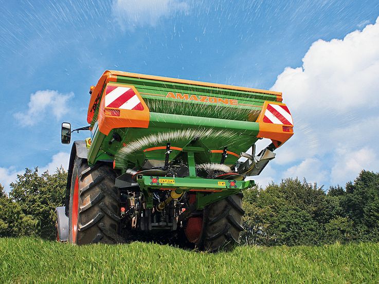 Un épandeur monté sur tracteur de couleurs verte et orange disperse du matériel sur un champ herbeux. L'épandeur est étiqueté « AMAZONE » et est entouré d'un ciel bleu avec des nuages en arrière-plan.