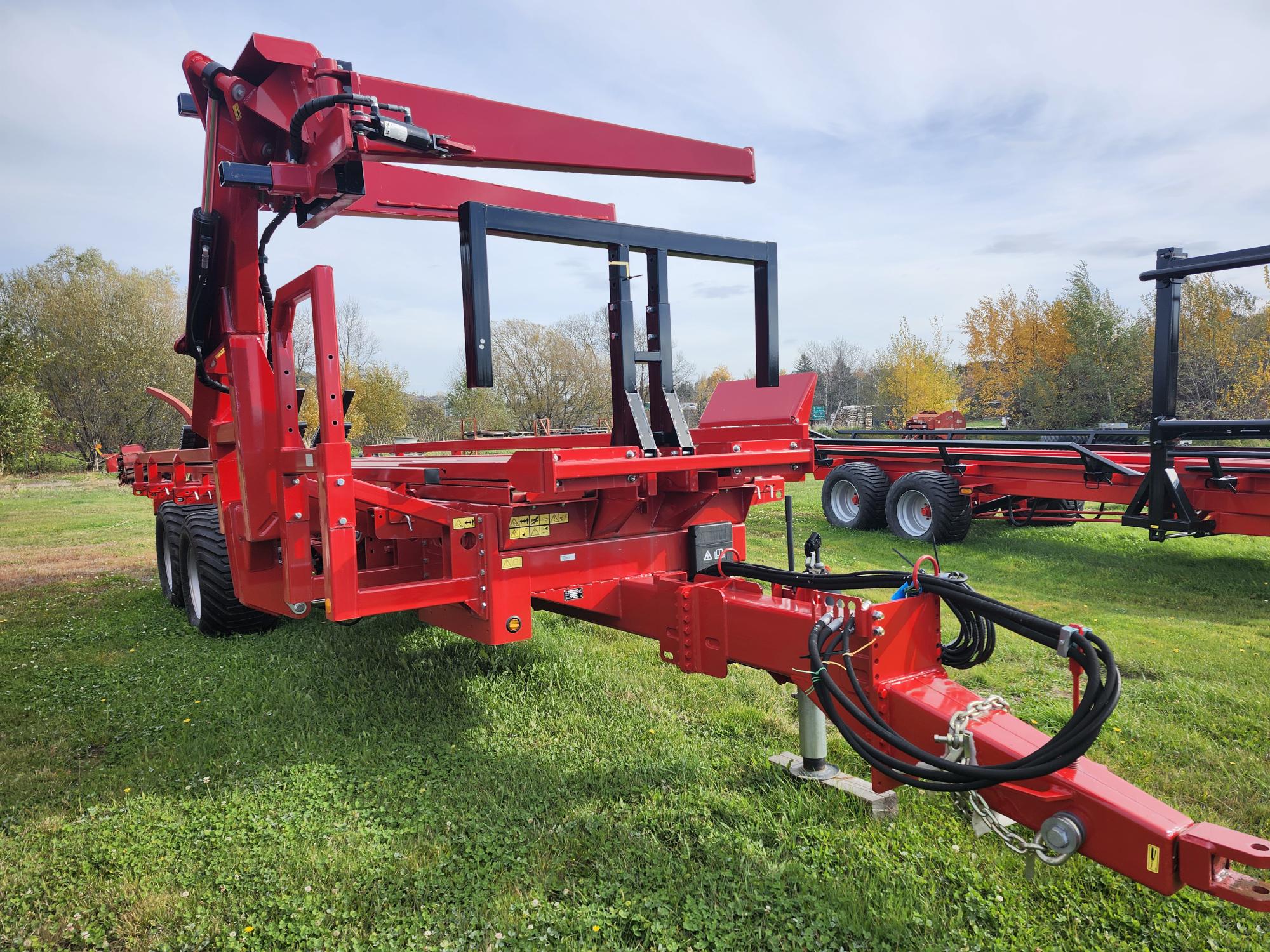 Des machines agricoles rouges sont posées sur un terrain herbeux, avec un grand châssis et un bras extensible. À l'arrière-plan, on voit d'autres pièces d'équipement similaires et des arbres sous un ciel partiellement nuageux.