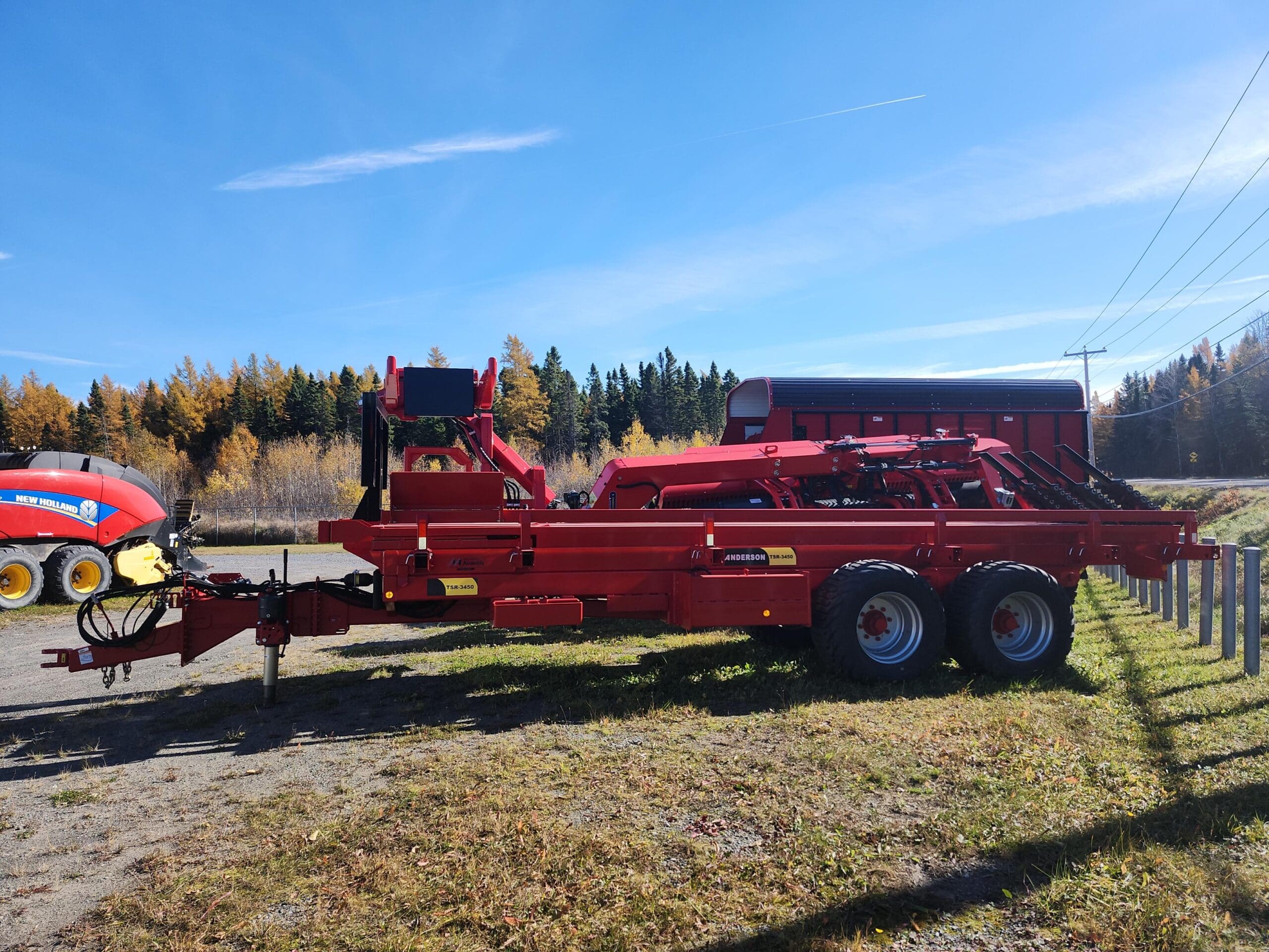 Une remorque agricole rouge est garée sur une zone herbeuse sous un ciel bleu clair. À l'arrière-plan, on voit des arbres au feuillage d'automne, une autre machine agricole rouge et une ligne électrique sur la droite.