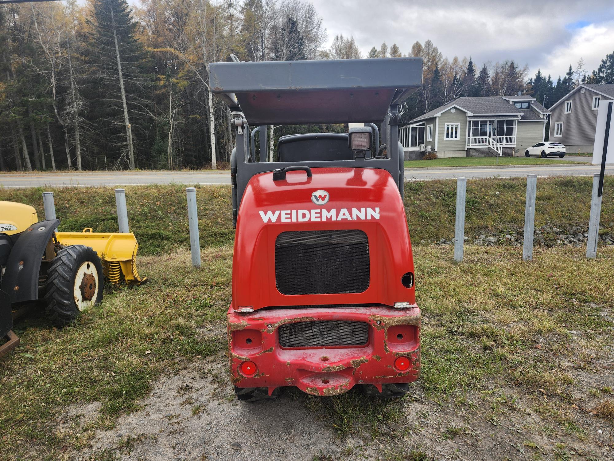 Un véhicule de chantier rouge de la marque Weidemann est garé sur un terrain herbeux avec des arbres en arrière-plan. Le véhicule est tourné vers l'extérieur et quelques maisons sont visibles au loin.