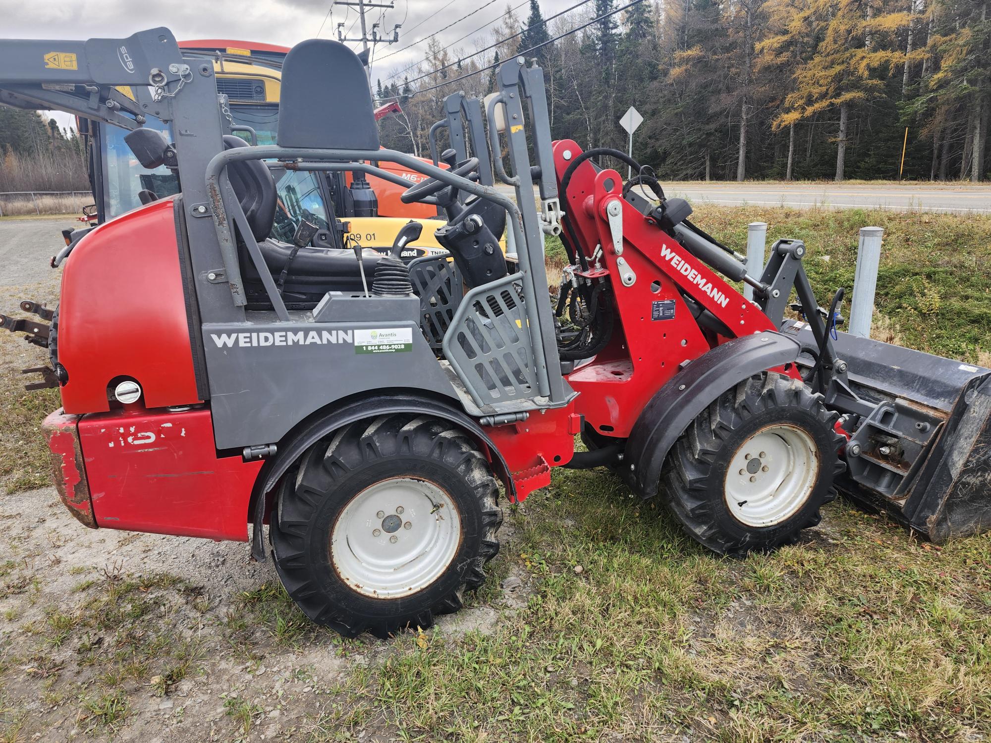 Une chargeuse compacte Weidemann rouge et grise est garée sur l'herbe au bord d'une route. La chargeuse est équipée de gros pneus et est orientée vers la gauche, avec un godet à l'avant. Des arbres et des poteaux électriques sont visibles à l'arrière-plan.