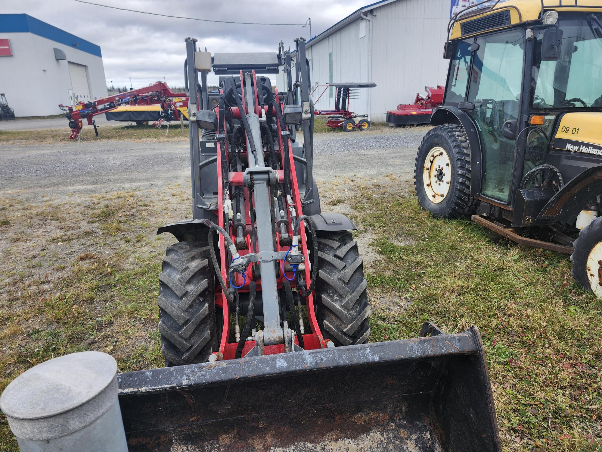 Un tracteur avec chargeur frontal est stationné sur l'herbe à côté d'un tracteur New Holland. Des équipements agricoles sont visibles en arrière-plan près d'un bâtiment industriel blanc. Ciel couvert au-dessus.