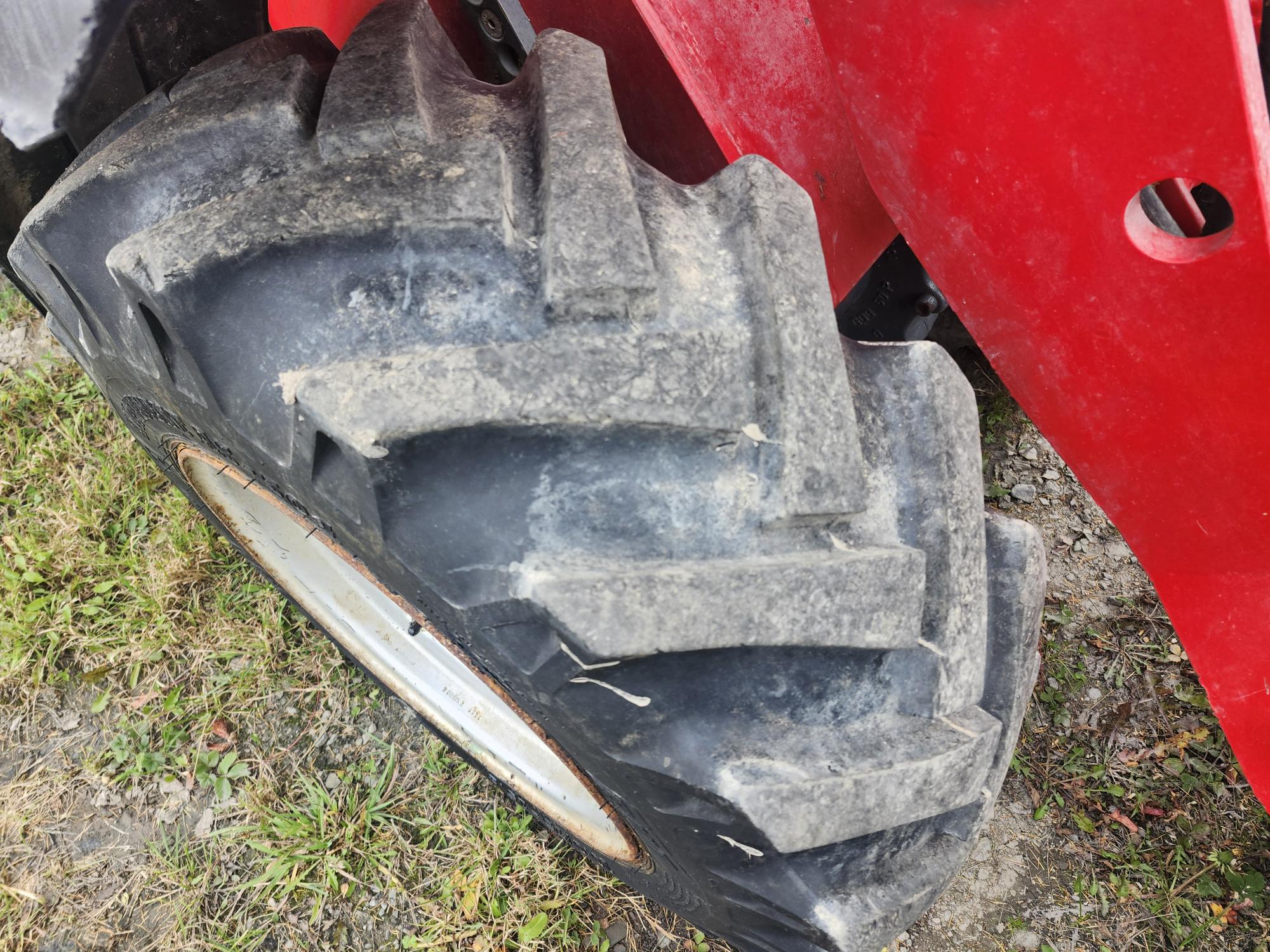 Gros plan d'un gros pneu de tracteur poussiéreux sur l'herbe. Le pneu est fixé à un véhicule rouge, peut-être un tracteur ou une machine agricole. Le dessin de la bande de roulement est visible et le décor semble être à l'extérieur sur une surface de gravier et d'herbe.