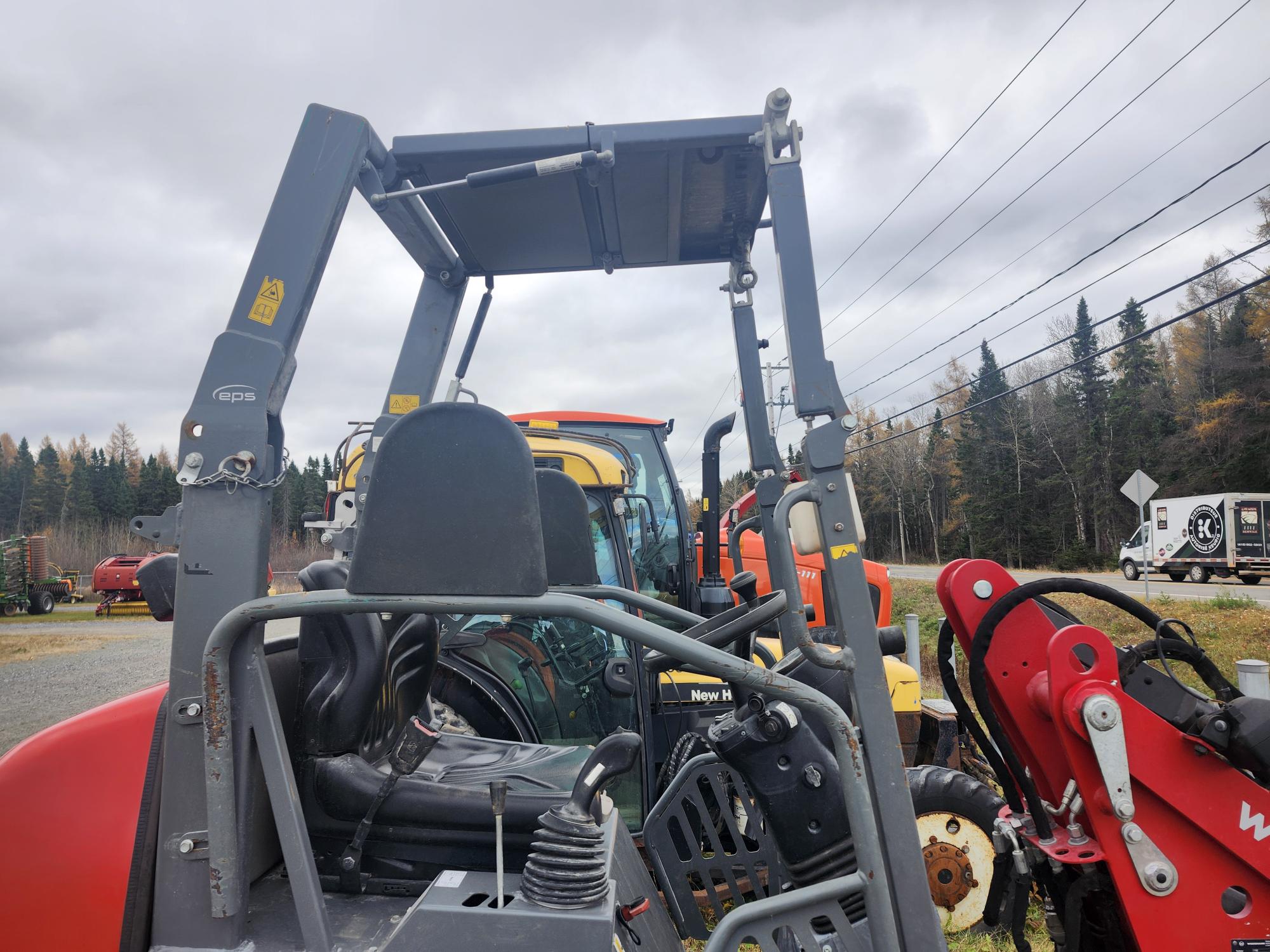 Gros plan sur un équipement agricole, notamment un bras de chargeur rouge et une cabine de tracteur avec un siège. À l'arrière-plan, on voit d'autres tracteurs et une zone boisée sous un ciel nuageux. Des lignes électriques traversent la scène.