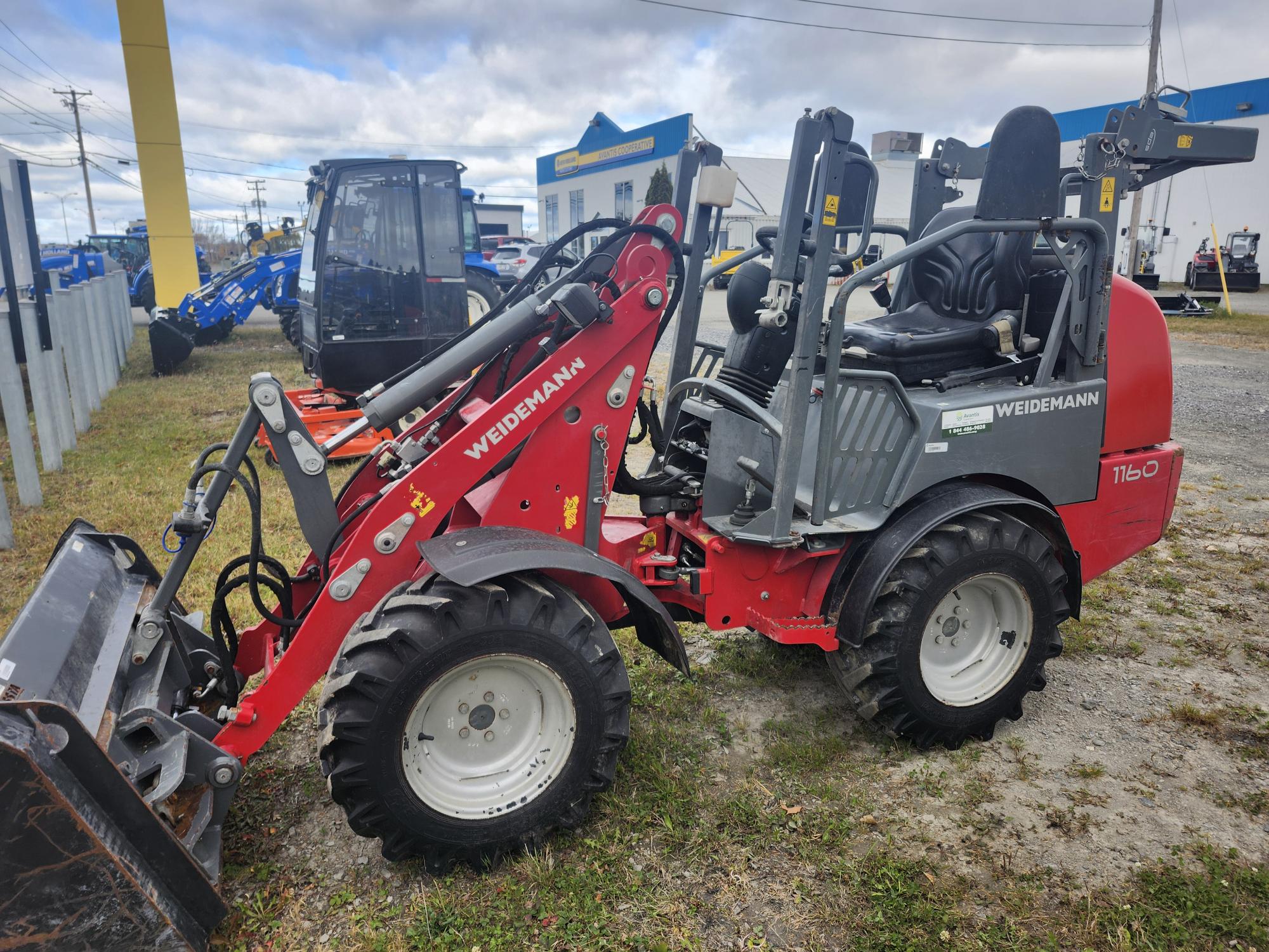 Un véhicule de chantier rouge de la marque Weidemann est garé sur une surface en gravier. Le véhicule est équipé de gros pneus, d'un chargeur frontal et d'une cabine ouverte. Des bâtiments et d'autres machines sont visibles à l'arrière-plan.