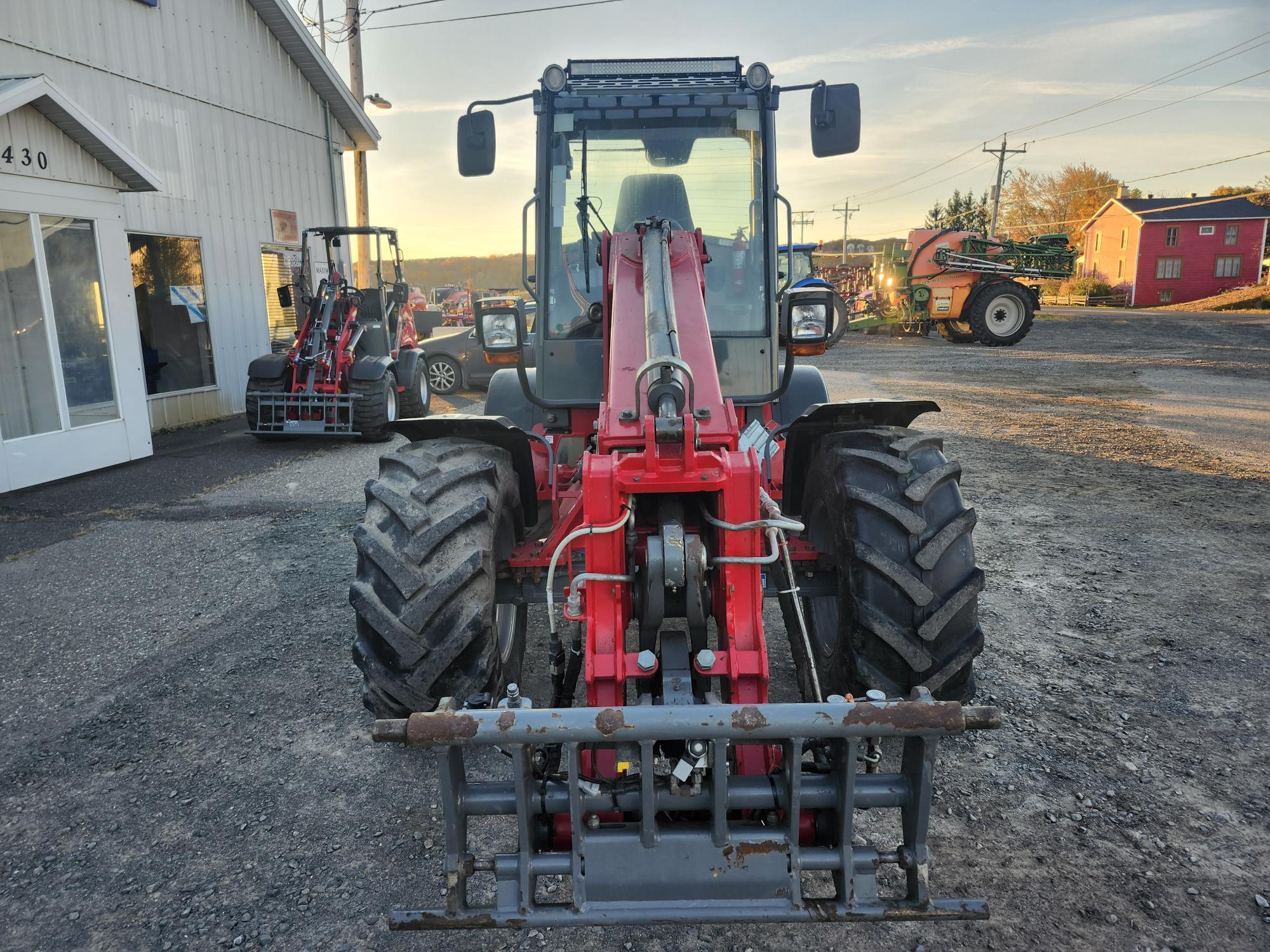 Un tracteur rouge avec de gros pneus noirs est garé sur un terrain en gravier. L'arrière-plan montre un autre véhicule similaire, un bâtiment et une grange rouge. Le ciel est clair, ce qui suggère une journée ensoleillée.