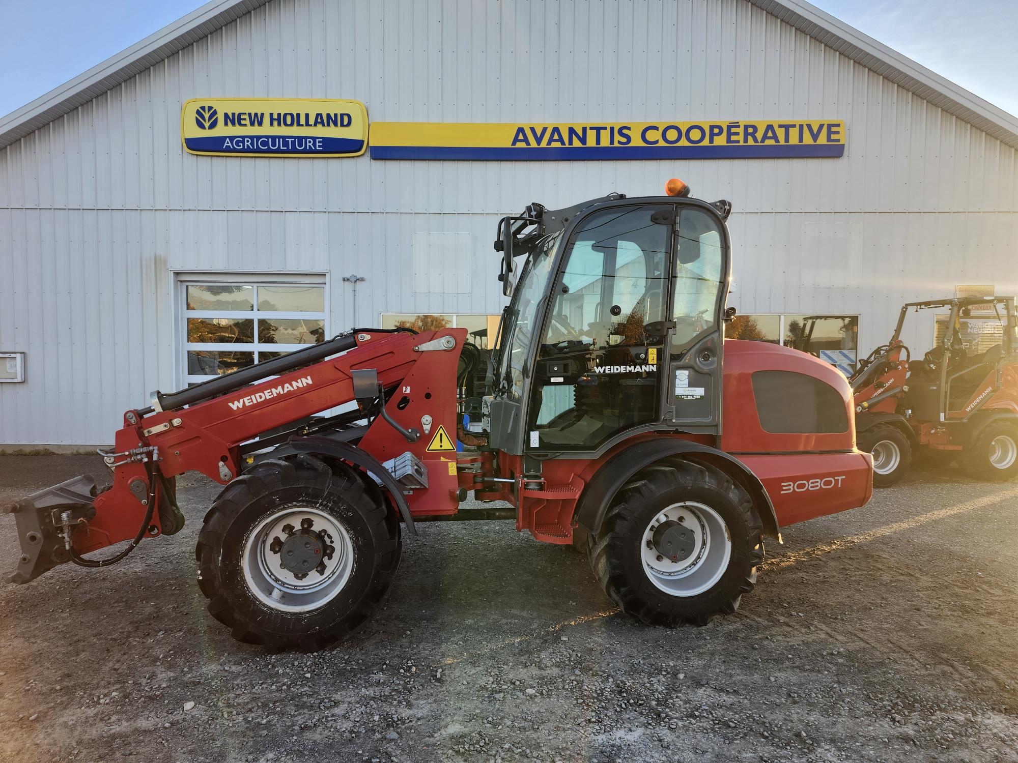 Un chariot télescopique rouge Weidemann 3080T est garé sur du gravier devant un bâtiment portant les inscriptions « New Holland Agriculture » et « Avantis Cooperative ». Un autre véhicule similaire est partiellement visible à l'arrière-plan.