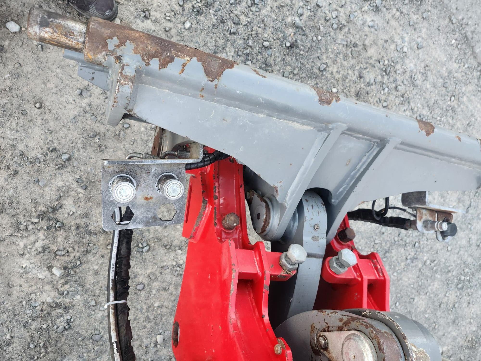 Gros plan d'un mécanisme industriel en métal posé sur une surface en béton brut. L'équipement est doté de composants rouges et gris avec des boulons, des fils et des taches de rouille visibles sur certaines surfaces.