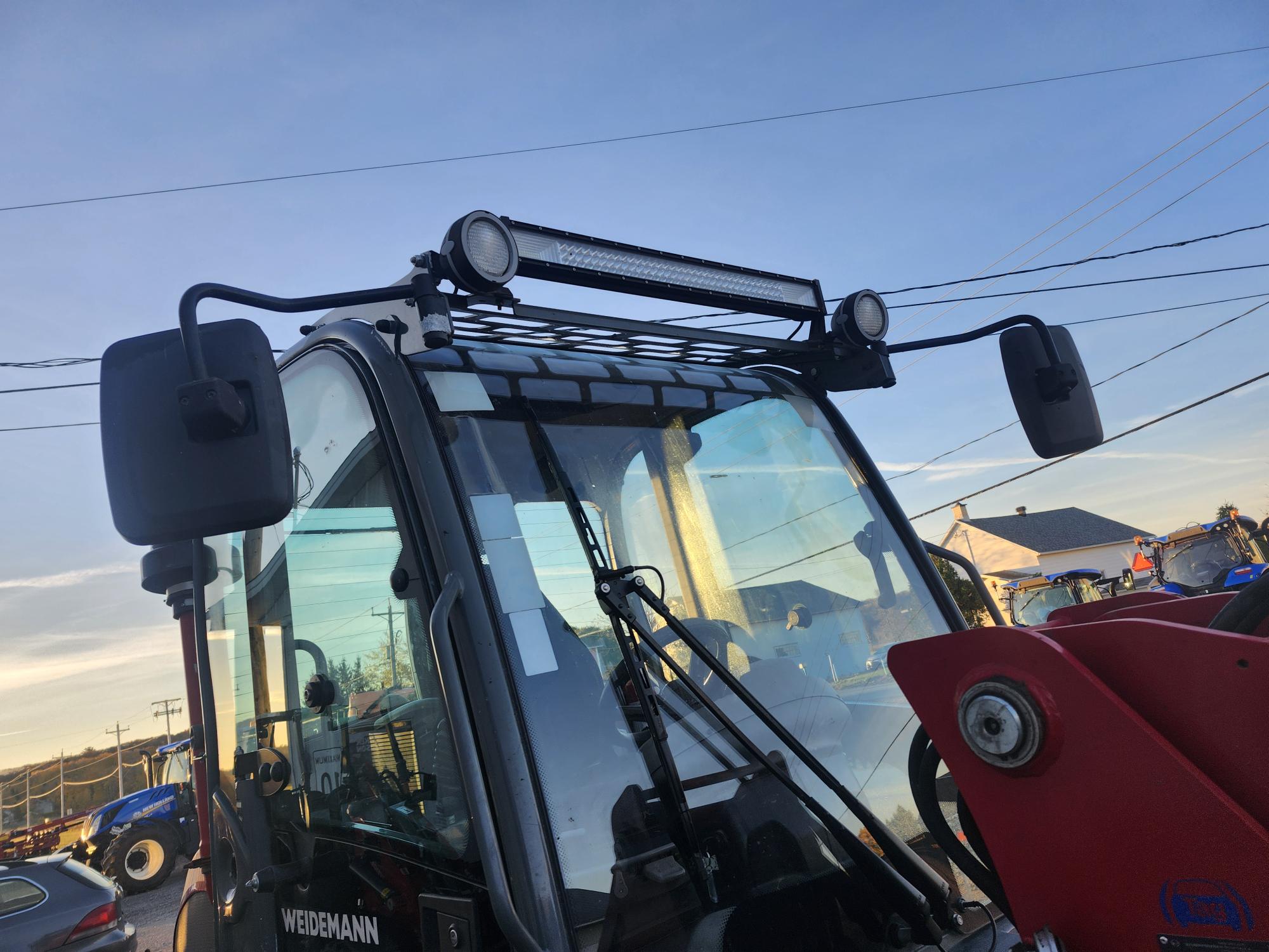 Gros plan d'un tracteur Weidemann rouge doté d'une cabine avec deux rétroviseurs latéraux et d'une barre lumineuse LED montée sur le toit. L'arrière-plan montre un ciel bleu clair, des fils électriques et d'autres équipements agricoles.