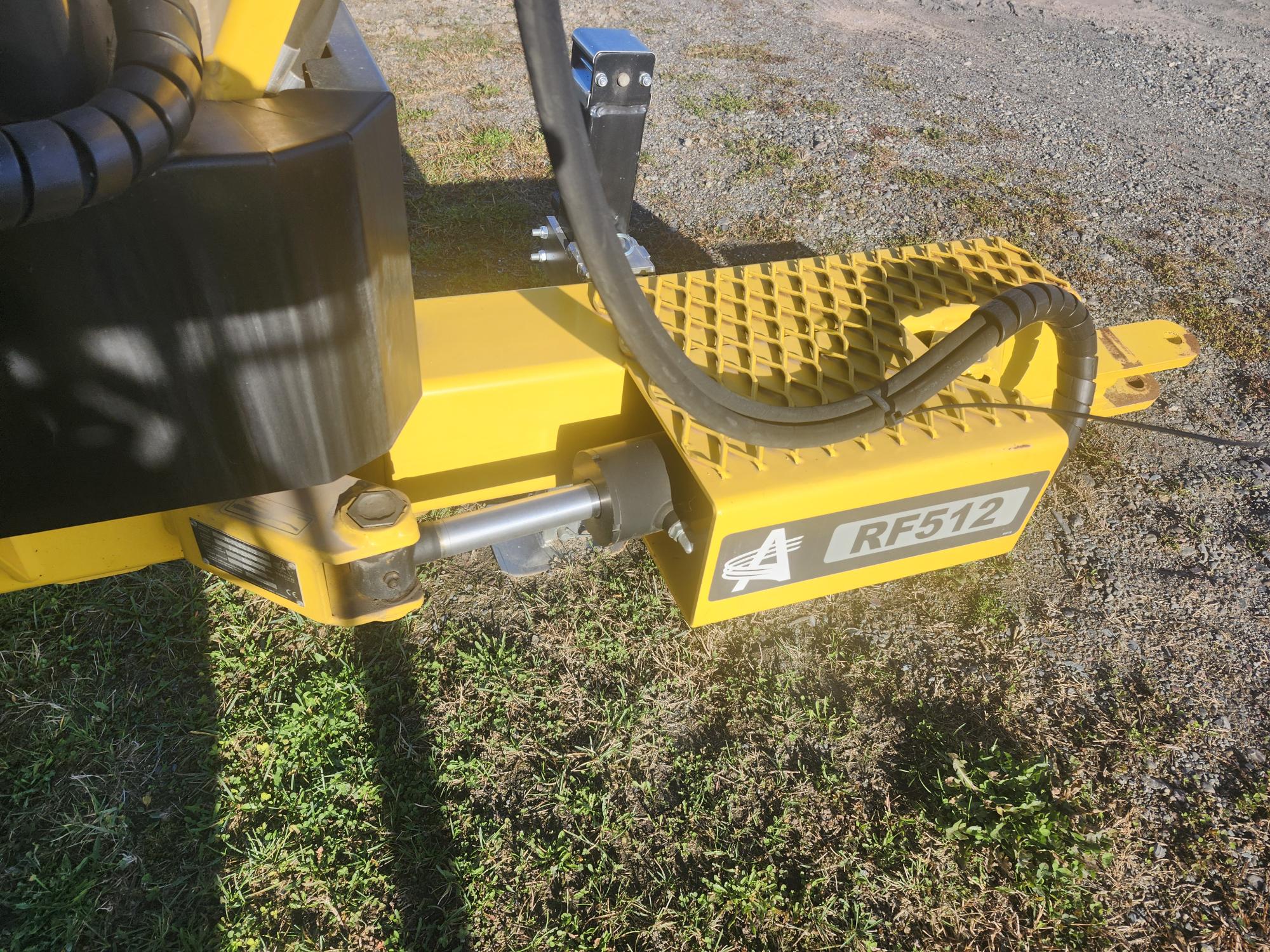 Gros plan d'un accessoire agricole jaune étiqueté « RF512 » avec une plaque de base à carreaux. L'accessoire est relié à une machine, installée sur une zone herbeuse et gravillonnée sous un soleil éclatant. D'épais câbles noirs sont visibles.