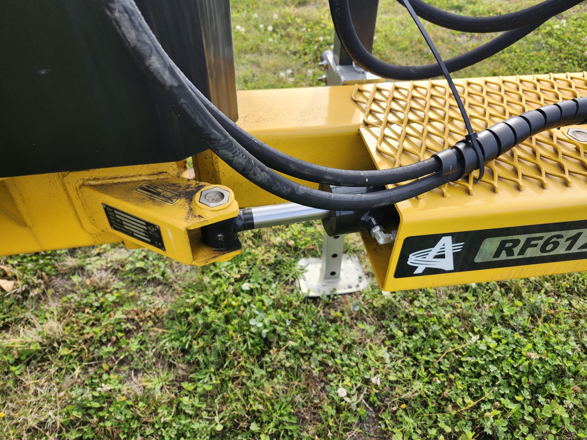 Gros plan d'un composant de machine industrielle jaune avec des tuyaux hydrauliques noirs et une étiquette marquée « RF612 ». La machine est placée sur l'herbe, avec un poteau de support métallique visible en dessous.