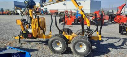 Une remorque de chargement de grumes jaune avec de grandes roues est garée sur un terrain en gravier, entourée de diverses autres machines. L'arrière-plan comprend un grand bâtiment industriel.