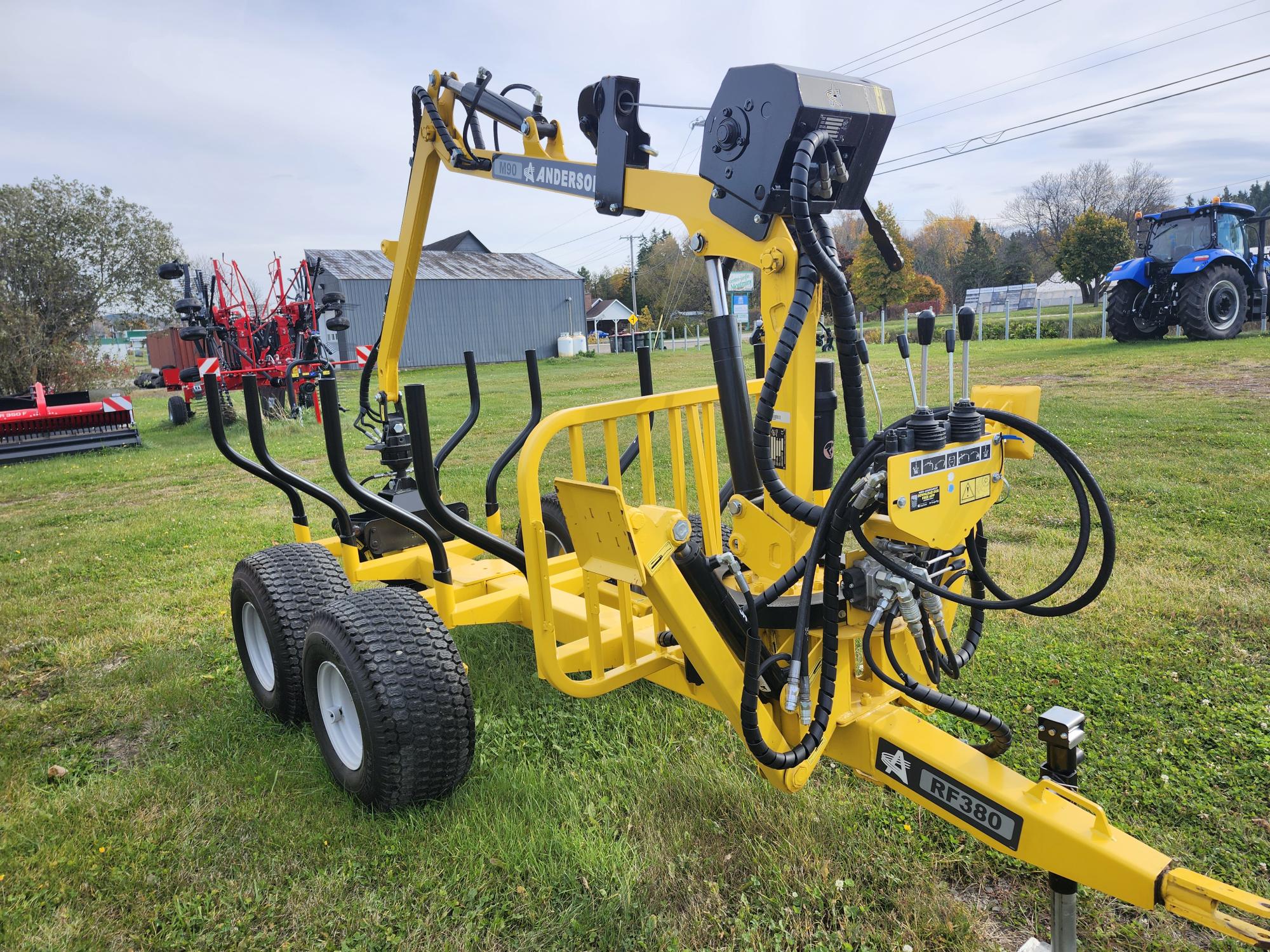 Une machine agricole jaune vif avec de gros pneus et divers composants hydrauliques se trouve sur un champ herbeux. À l'arrière-plan, on voit des bâtiments agricoles, des arbres et d'autres équipements agricoles.