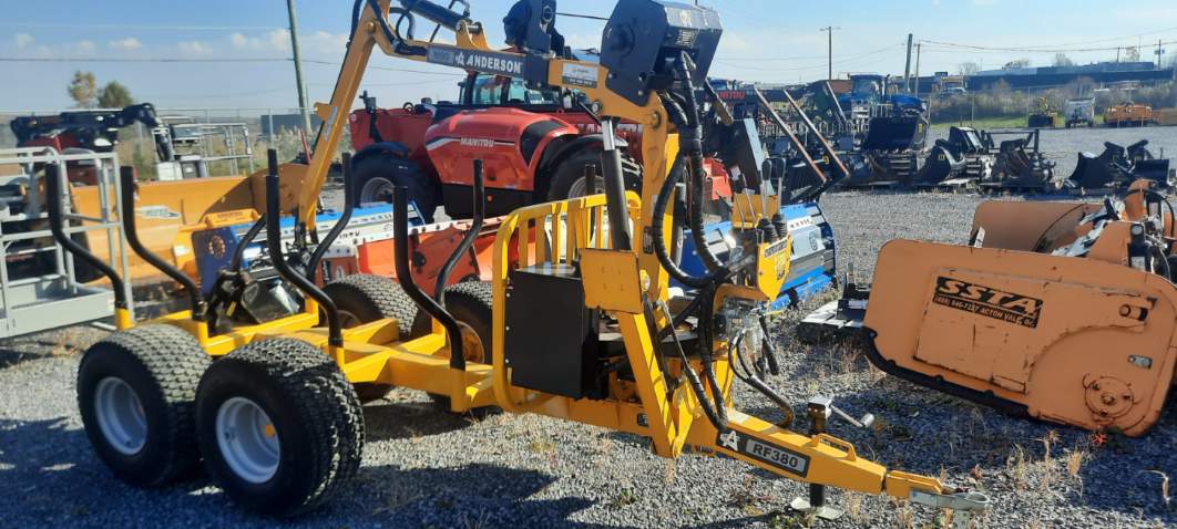 Une enrubanneuse Anderson jaune garée sur du gravier avec diverses machines agricoles en arrière-plan. Elle est équipée de roues jumelées et de composants hydrauliques visibles.