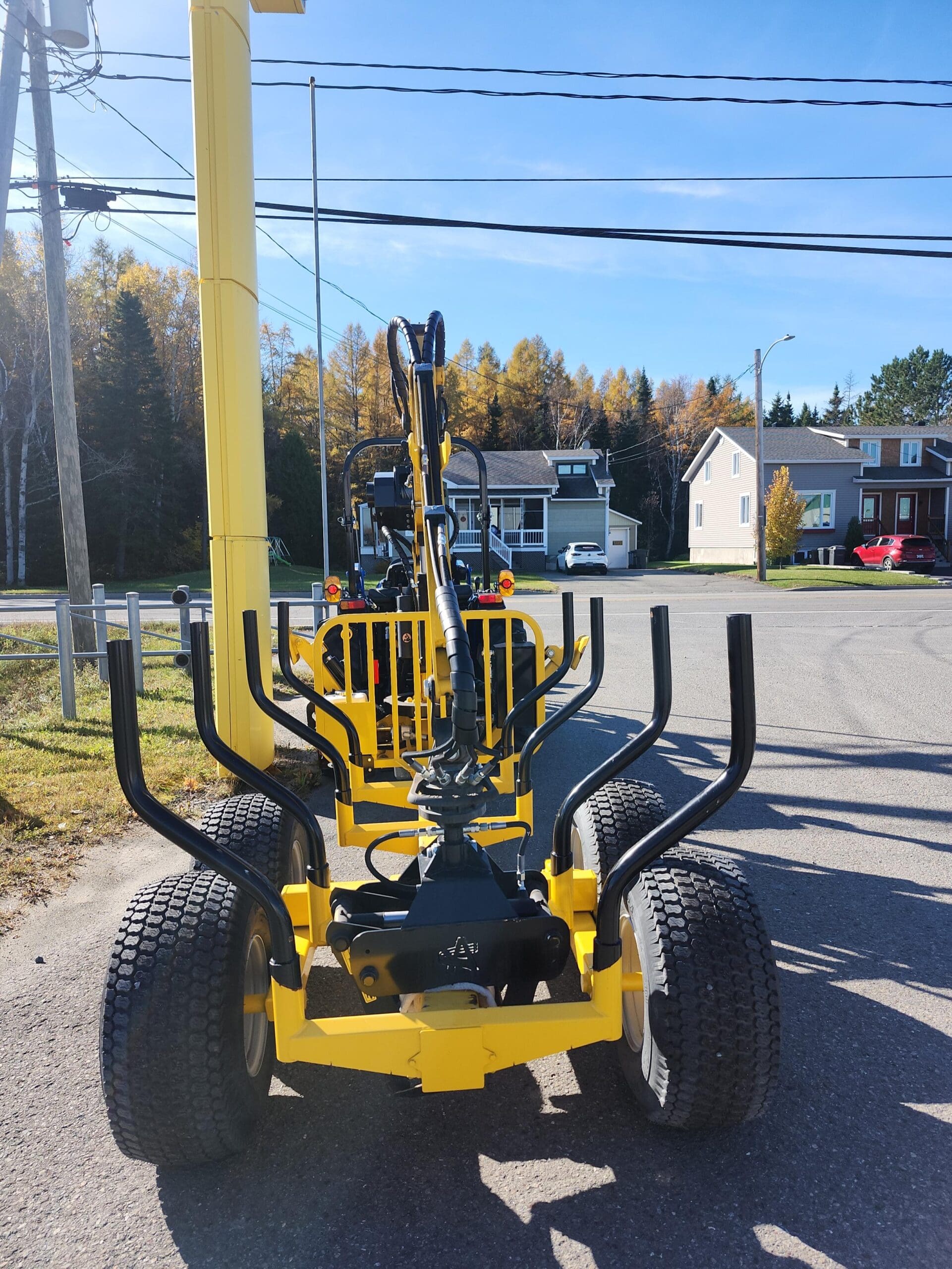 Un accessoire de machinerie jaune pour un tracteur, équipé d'un grappin, est garé sur une route pavée. À l'arrière-plan, on voit des maisons et des arbres au feuillage d'automne sous un ciel bleu clair.