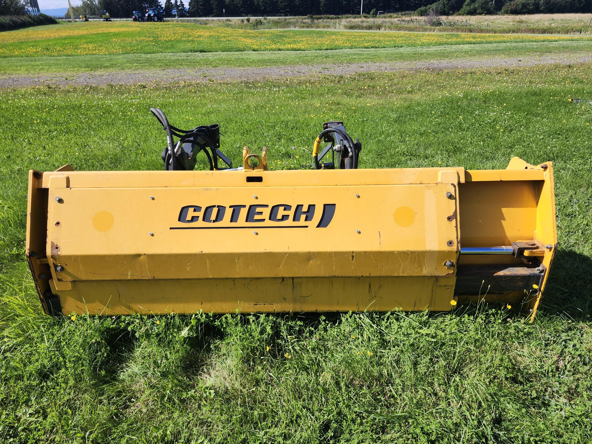 Une machine industrielle jaune étiquetée « COTECH » est exposée sur un sol herbeux. Elle est installée à l'extérieur avec un champ et des arbres en arrière-plan sous un ciel bleu clair.