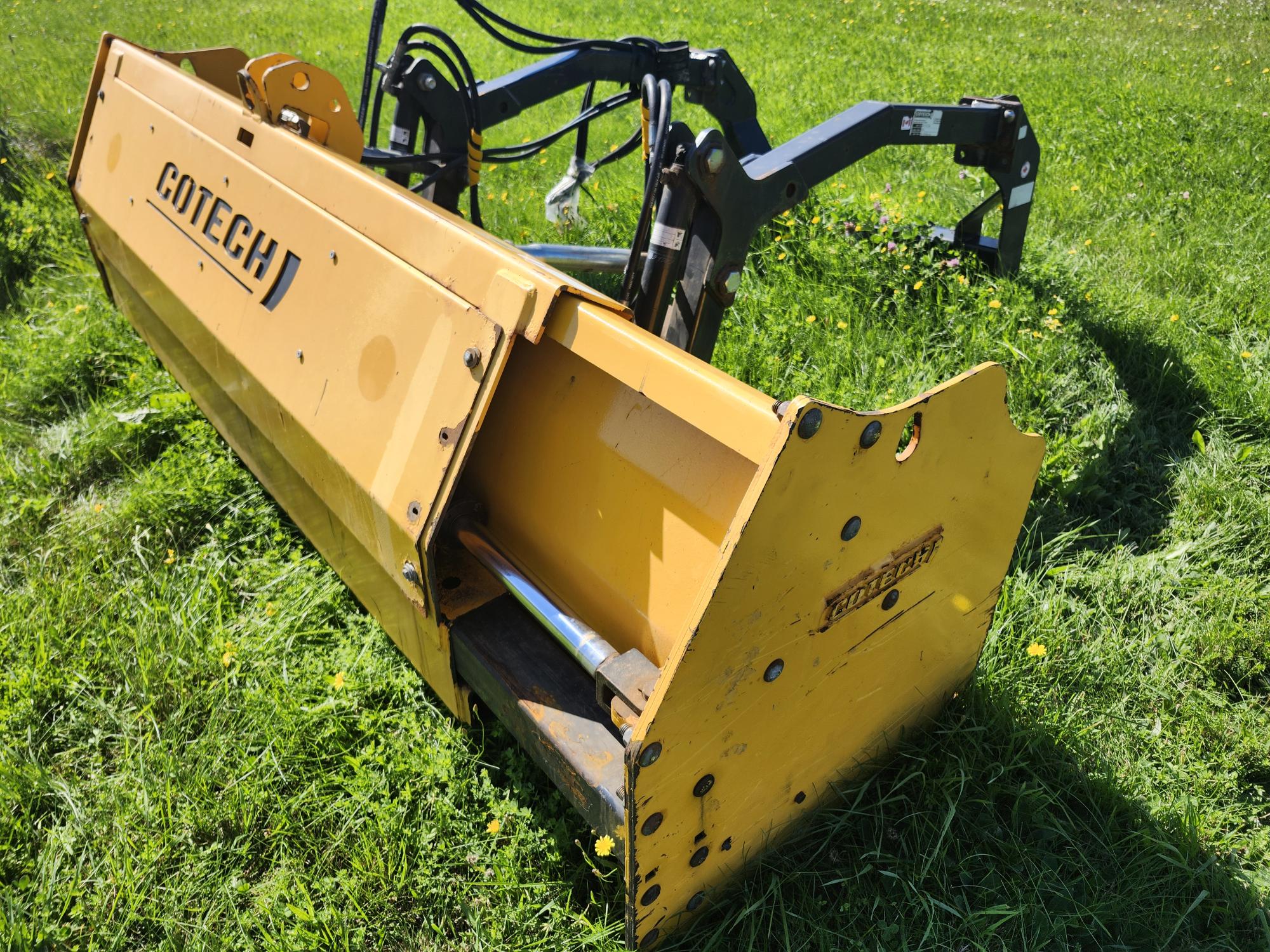 Accessoire de niveleuse jaune sur un terrain herbeux sous le soleil, doté de composants hydrauliques et de la marque « Cotech » sur le côté.