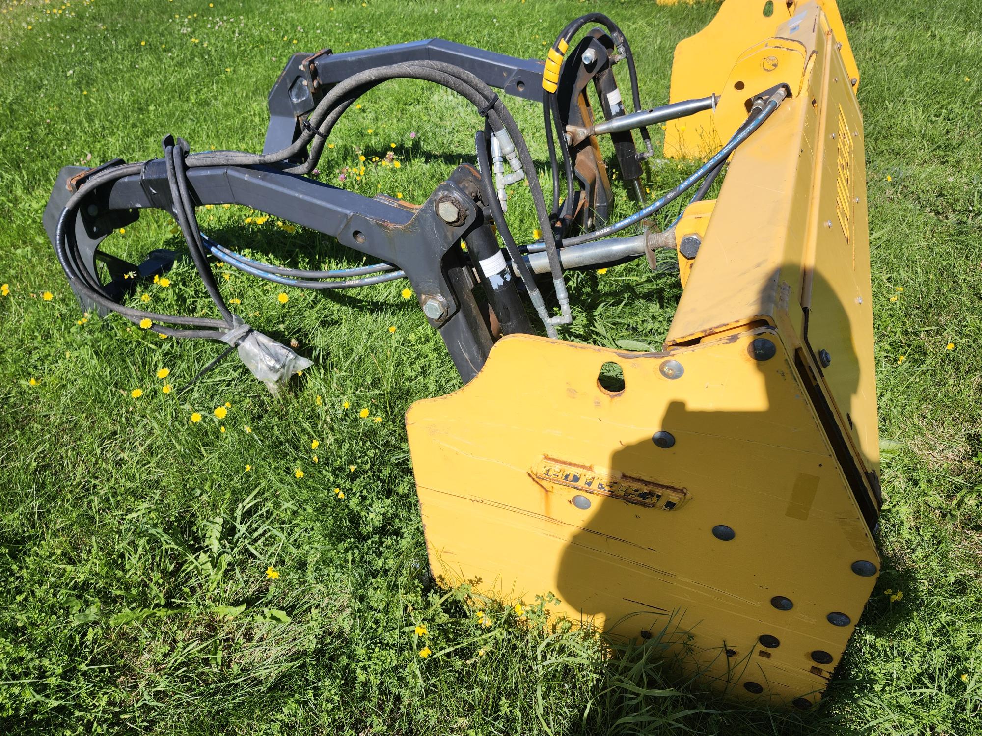 Une machine lourde jaune et noire est posée sur un terrain herbeux parsemé de petites fleurs jaunes. La machine est équipée de bras et de connexions hydrauliques et projette une ombre sur l'herbe.