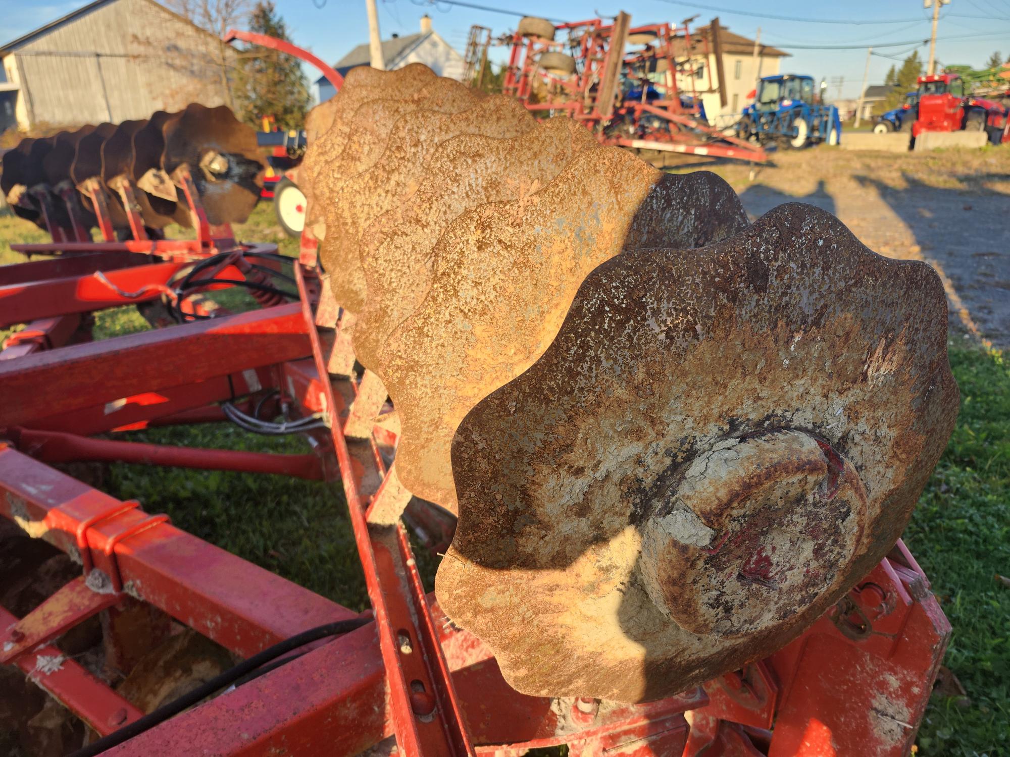 Gros plan d'un ensemble de disques de charrue circulaires rouillés fixés à un outil agricole rouge. L'arrière-plan montre d'autres équipements agricoles, une grange et une zone herbeuse sous un ciel clair.