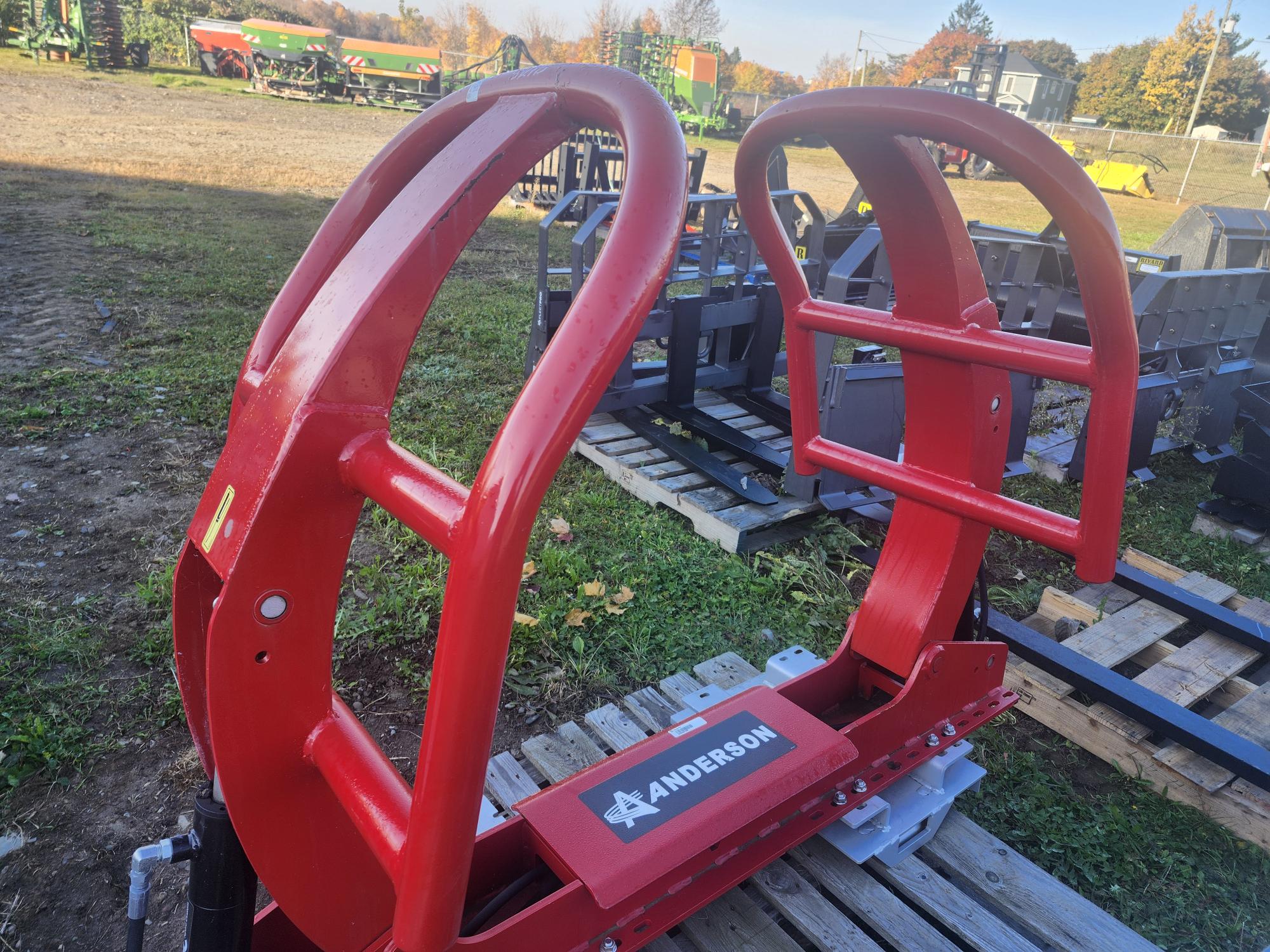 Une manutentionneuse de balles agricoles Anderson rouge placée sur des palettes en bois à l'extérieur, avec divers équipements agricoles visibles en arrière-plan sous un ciel clair.