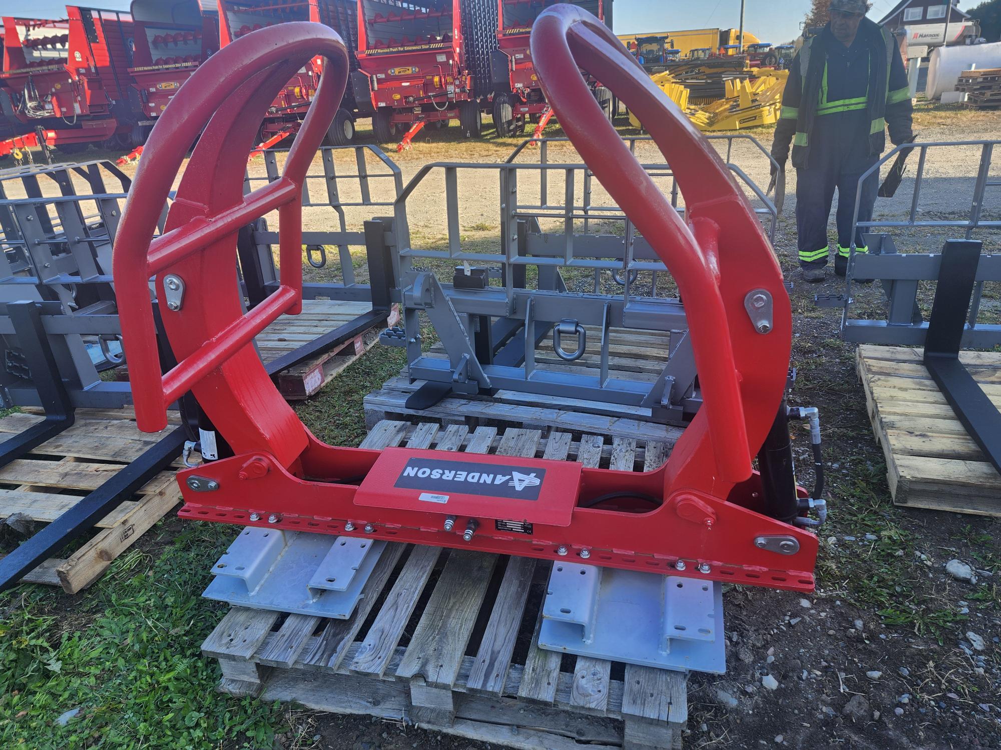 Un équipement lourd rouge est exposé à l'extérieur sur des palettes en bois. À l'arrière-plan, on voit d'autres composants de machines et une personne portant des vêtements de travail réfléchissants.