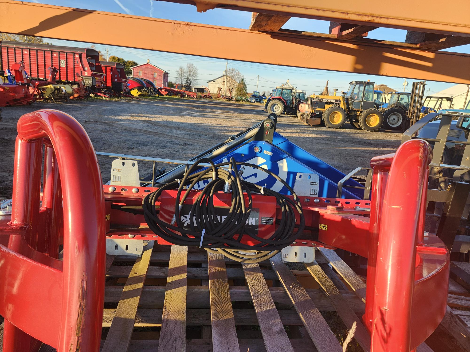 Un accessoire agricole rouge équipé de câbles hydrauliques est placé sur une palette en bois. À l'arrière-plan, diverses machines et équipements agricoles, ainsi que des bâtiments et un tracteur, sont visibles sous un ciel clair.