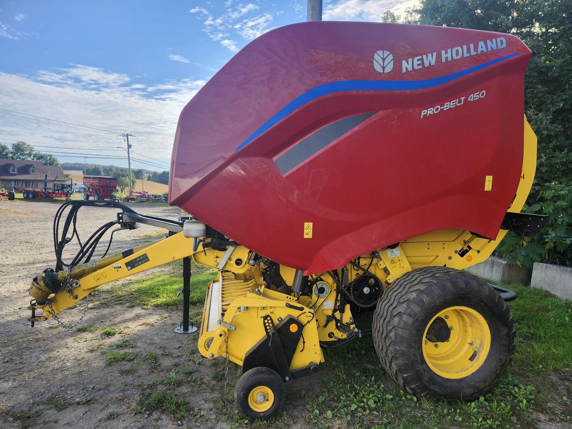 Une presse à balles rondes New Holland Pro-Belt 450 rouge et jaune est garée sur une surface en terre. Des arbres et un bâtiment sont visibles en arrière-plan sous un ciel partiellement nuageux.