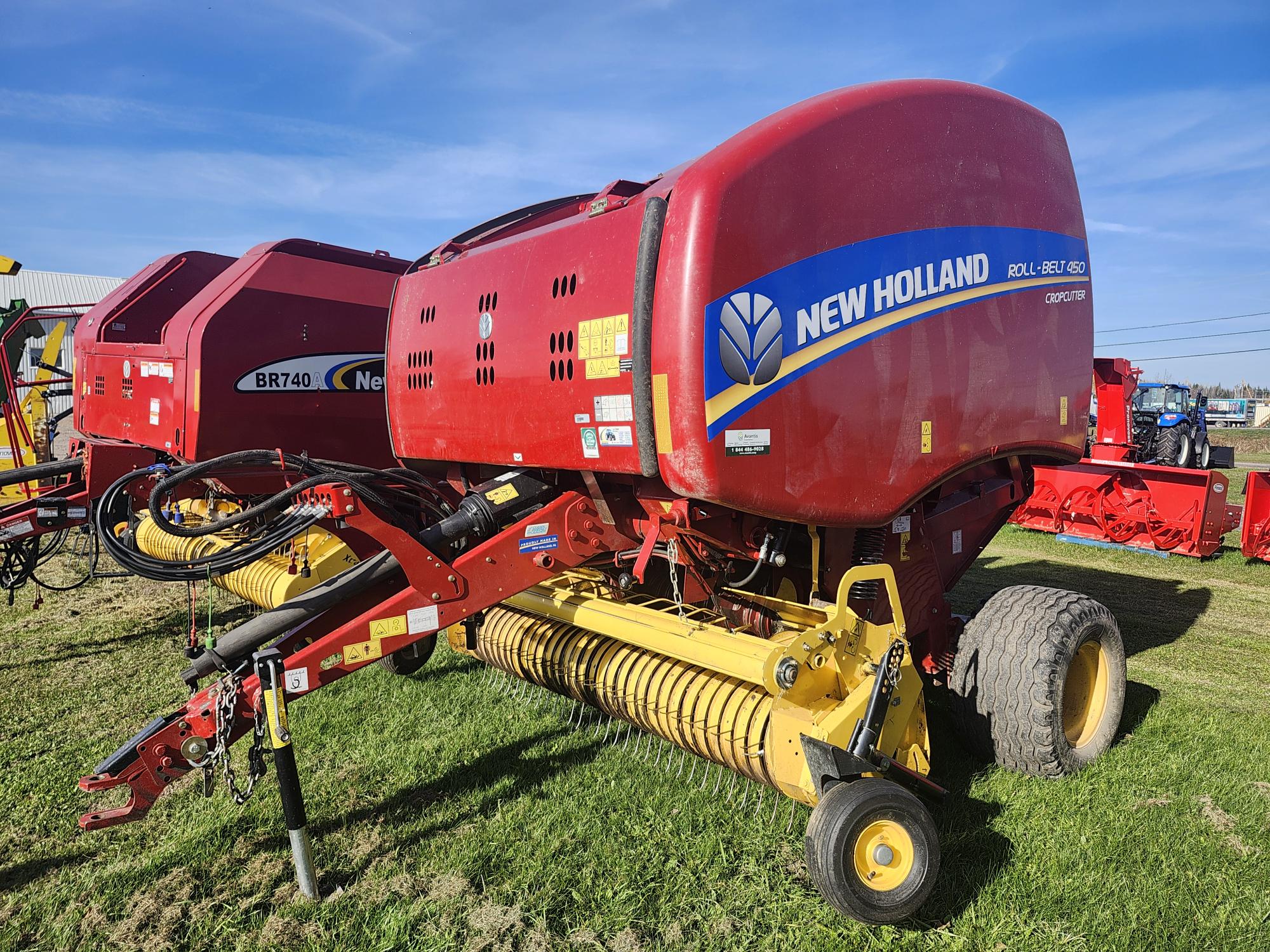 Une presse à balles rondes New Holland rouge garée sur l'herbe sous un ciel bleu clair. La machine est équipée d'un pick-up jaune, de roues et d'autocollants de marque. D'autres équipements agricoles sont visibles en arrière-plan.