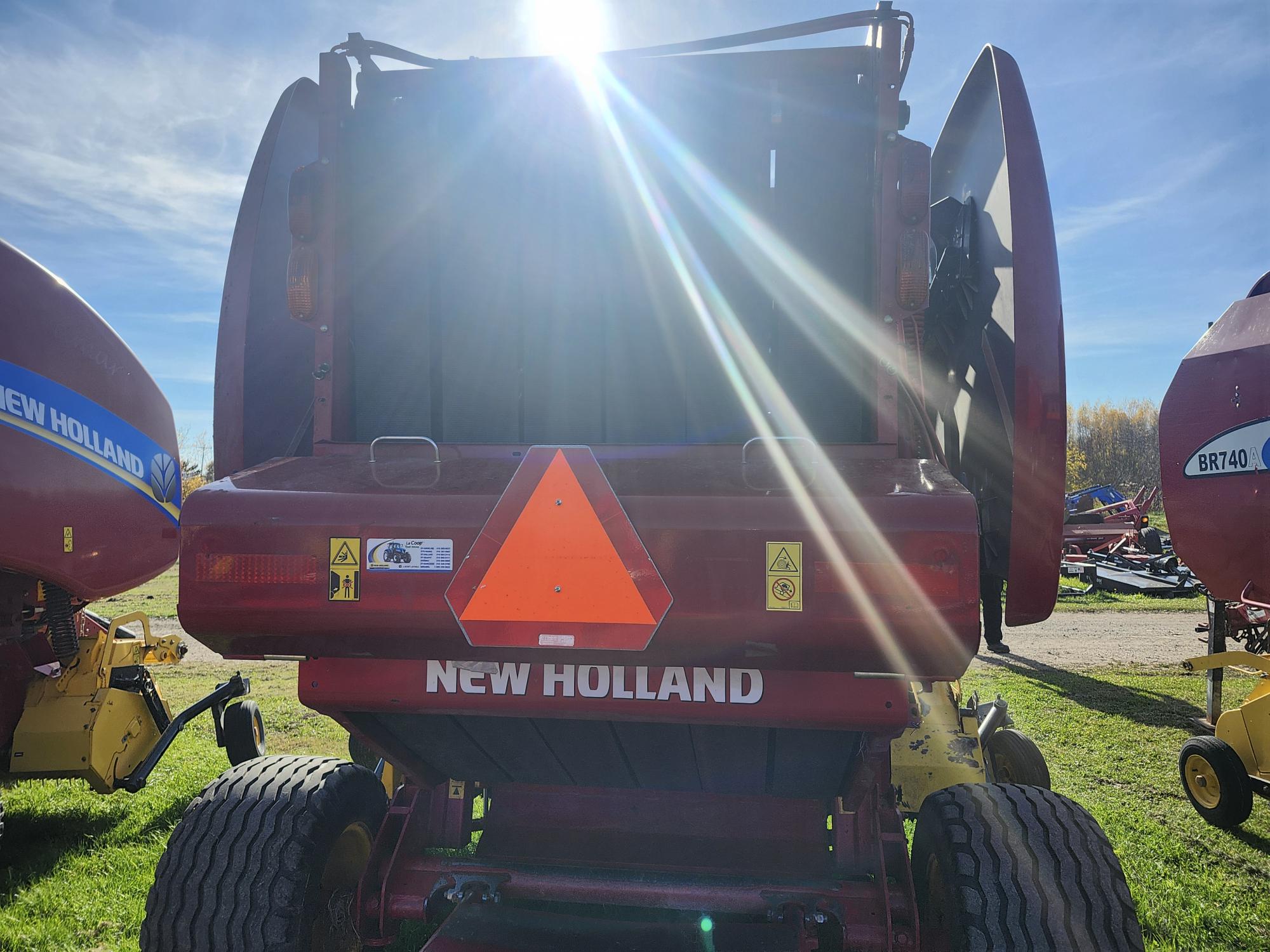 Une presse à balles New Holland rouge est garée sur l'herbe sous un ciel bleu clair. Le soleil brille fort d'en haut, projetant des rayons sur la machine. Un grand triangle d'avertissement orange est fixé à l'arrière. D'autres équipements agricoles sont visibles à l'arrière.