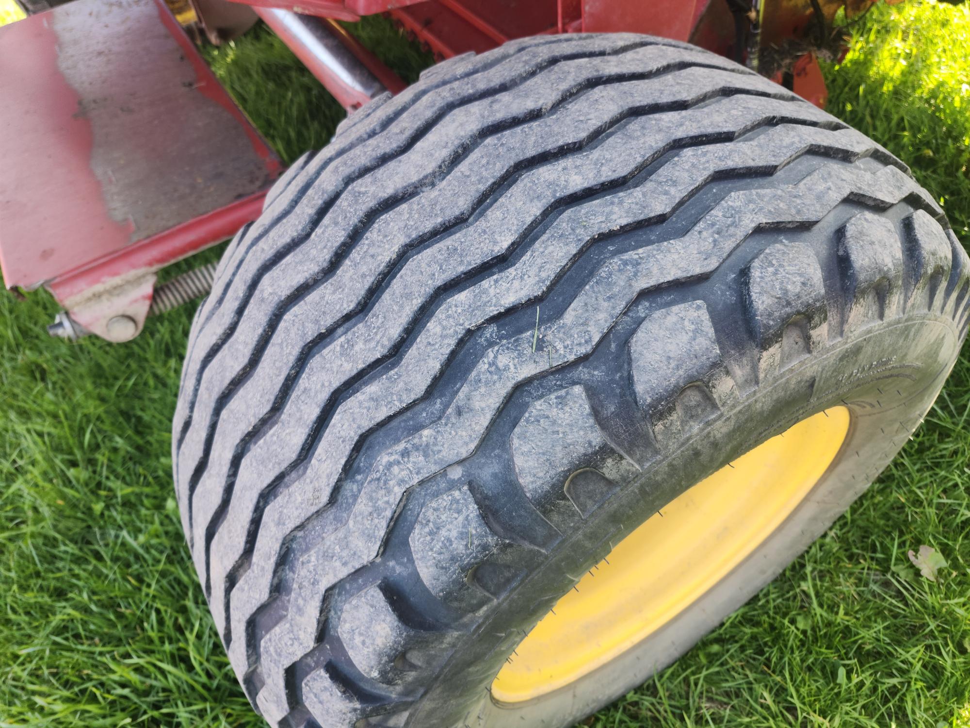 Gros plan d'une roue de tracteur avec une jante jaune et une bande de roulement texturée, posée sur une herbe verte luxuriante. La carrosserie rouge du tracteur est partiellement visible à l'arrière-plan.