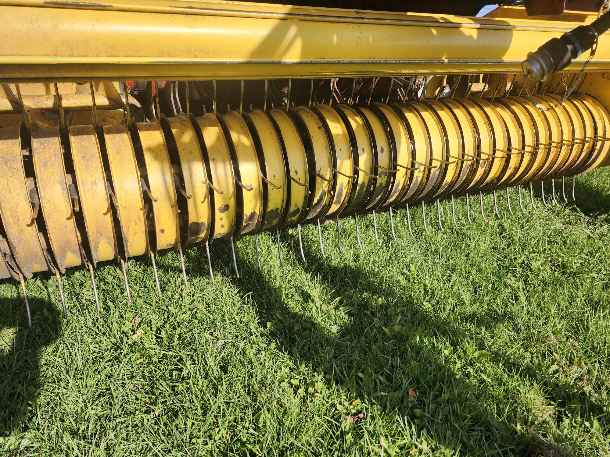 Gros plan d'une machine agricole jaune à dents rotatives, conçue pour la récolte des récoltes. Elle est positionnée au-dessus d'un champ d'herbe verte, projetant des ombres sur le sol.