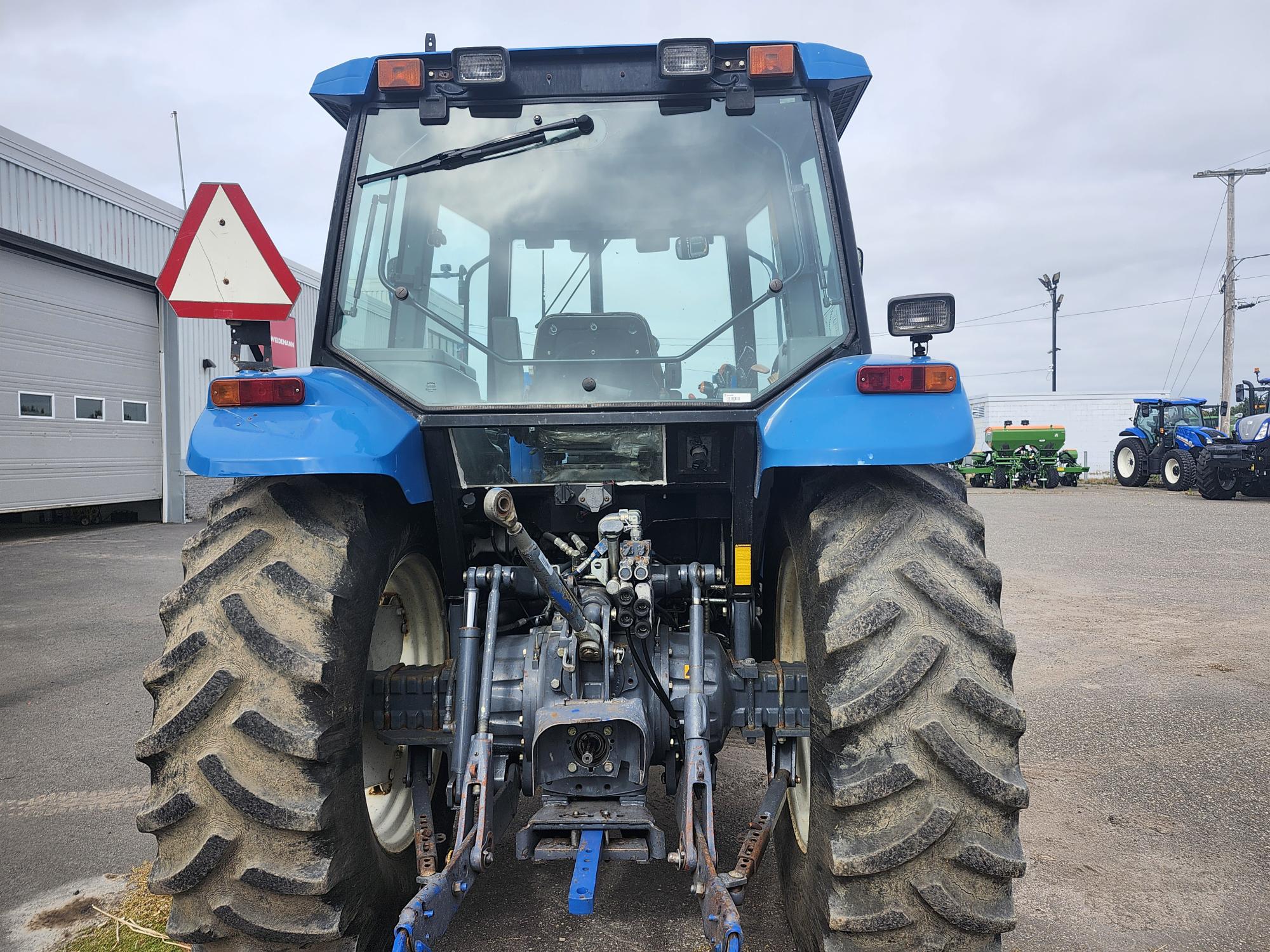 L'image montre la vue arrière d'un tracteur bleu stationné à l'extérieur. Il est doté de gros pneus robustes et de pièces de machinerie visibles. D'autres tracteurs et équipements agricoles sont placés à l'arrière-plan près d'un entrepôt. Le ciel est couvert.