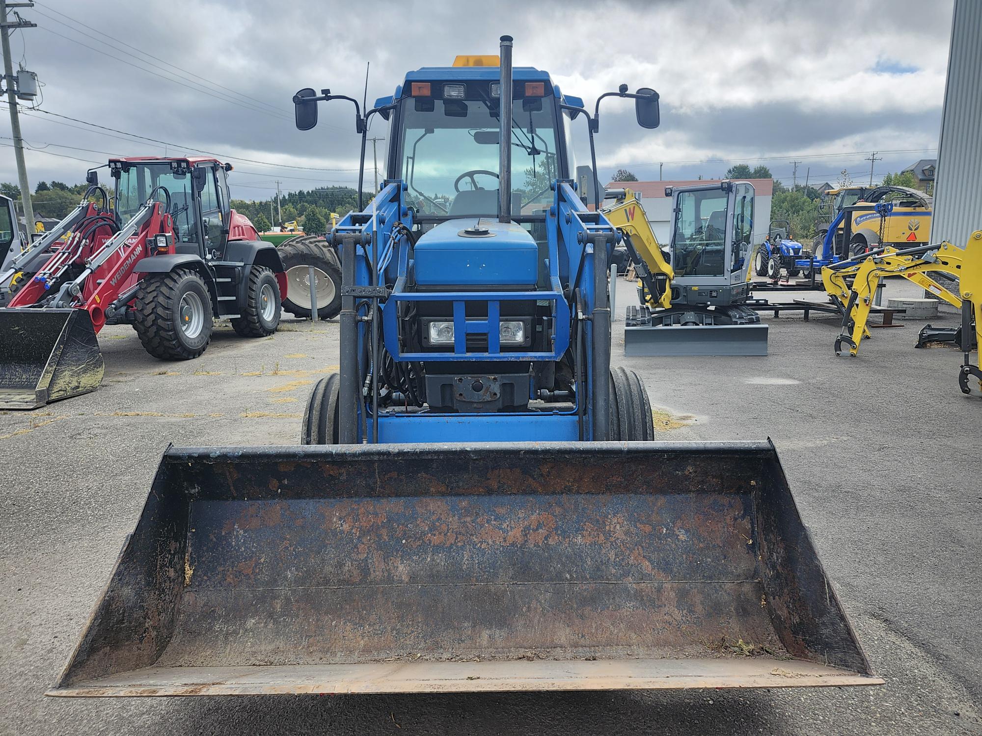 Un tracteur bleu à chargeur frontal équipé d'un gros godet est stationné sur un terrain extérieur entouré de machines lourdes, dont un tracteur rouge et une excavatrice jaune. Le ciel est nuageux et le sol est pavé d'asphalte.