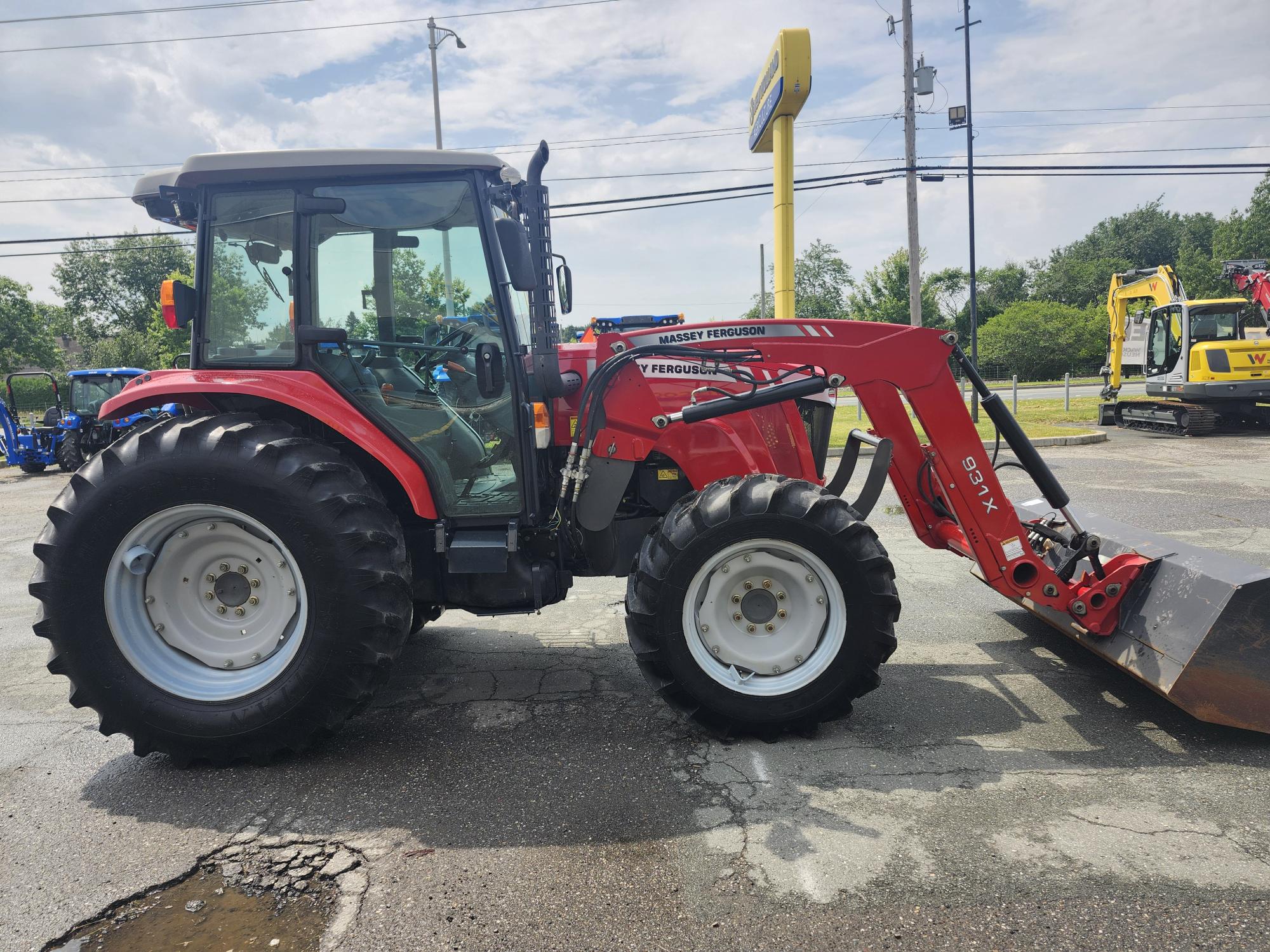 Un tracteur rouge Massey Ferguson équipé d'un chargeur frontal est stationné sur le trottoir. D'autres machines et un panneau sont visibles à l'arrière-plan. Le ciel est partiellement nuageux.