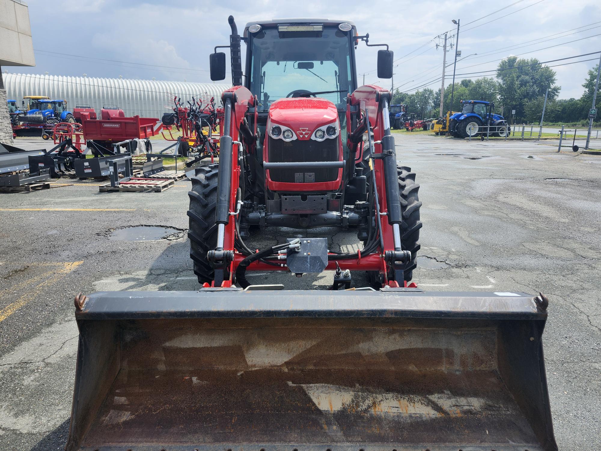 Un tracteur rouge avec un godet chargeur frontal est stationné sur une surface asphaltée. Plusieurs autres tracteurs et équipements agricoles sont visibles en arrière-plan sous un ciel partiellement nuageux.