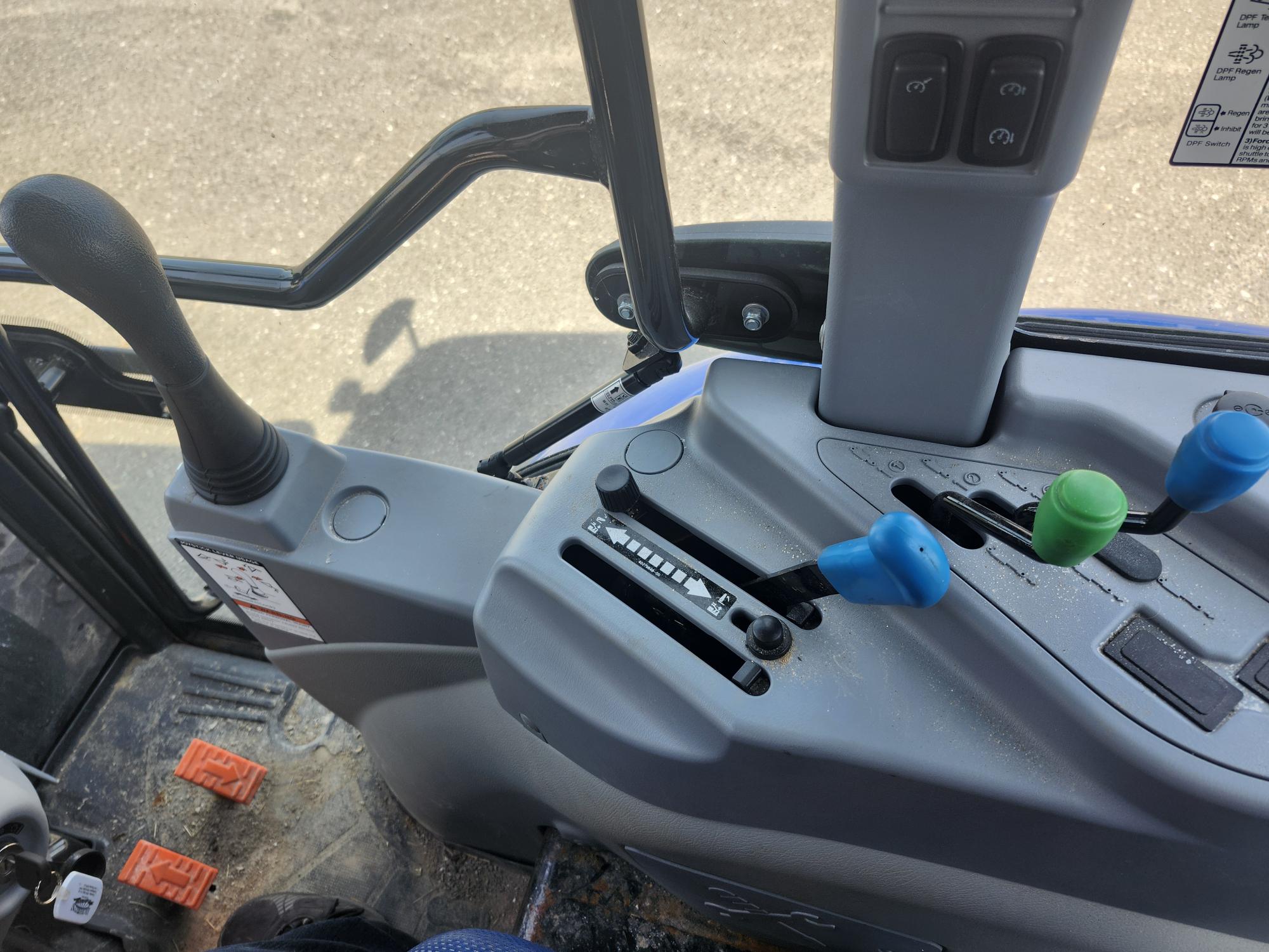 Vue du panneau de commande intérieur d'un tracteur avec divers leviers et interrupteurs. Le panneau comprend des leviers noirs, bleus et verts, ainsi que deux pédales rouges posées au sol sur un fond de trottoir.