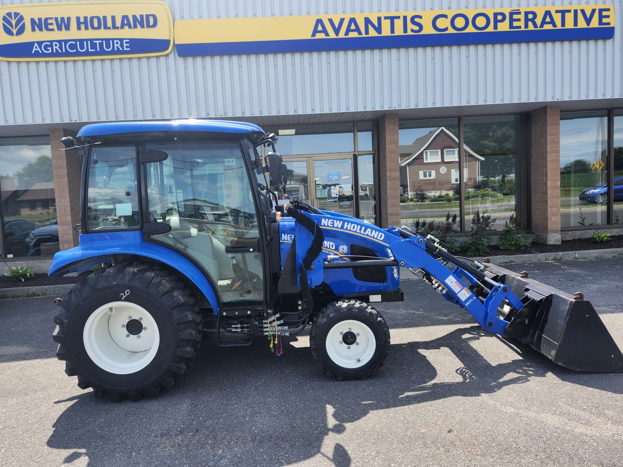 Un tracteur New Holland bleu équipé d'un chargeur frontal est garé devant une concession New Holland Agriculture avec un panneau indiquant la coopérative Avantis au-dessus de l'entrée. La scène se déroule par une journée ensoleillée.