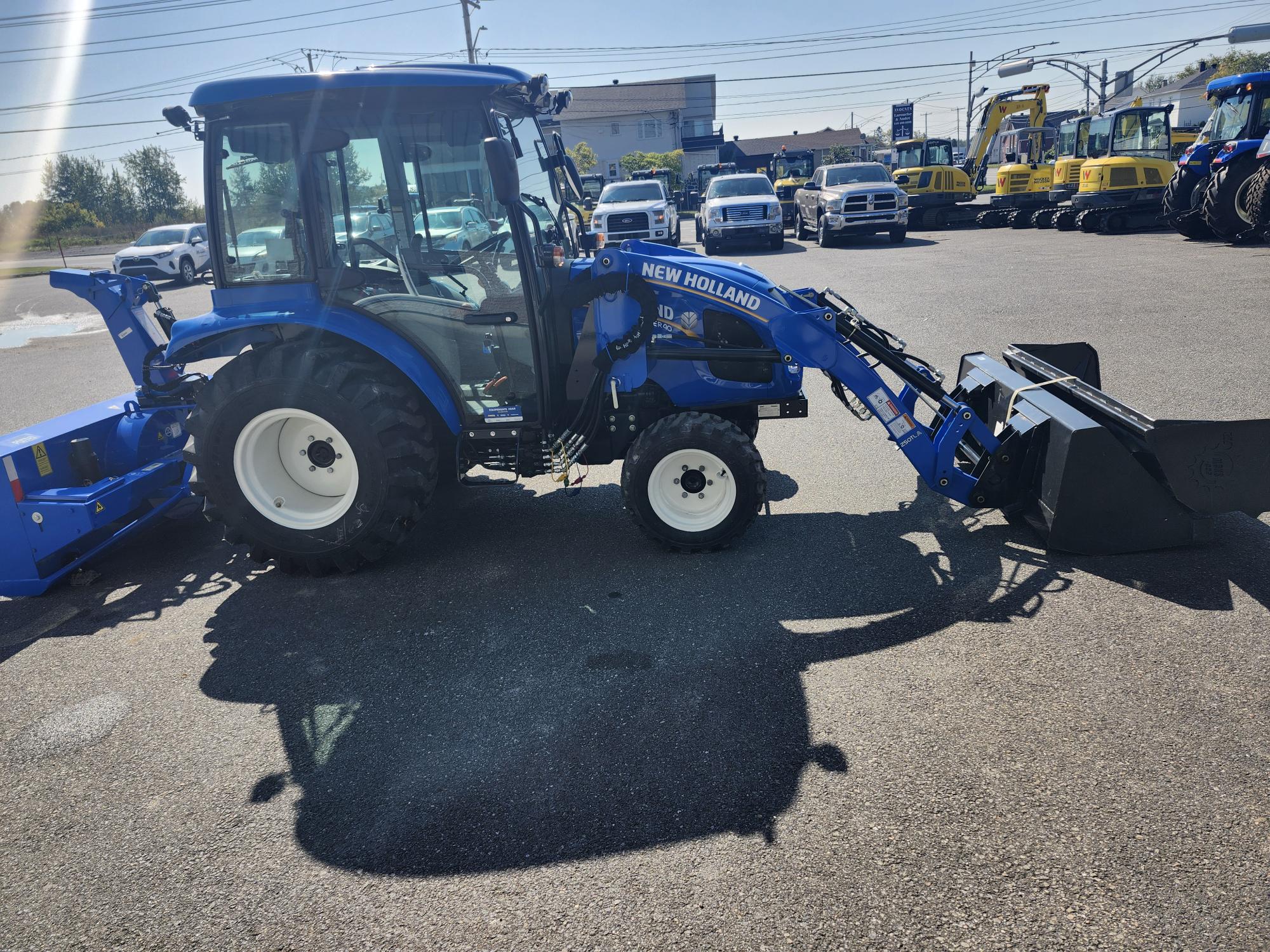 Un tracteur New Holland bleu équipé d'un chargeur frontal est garé sur un terrain pavé, entouré d'autres machines agricoles. Le soleil projette des ombres nettes, signe d'une journée ensoleillée.