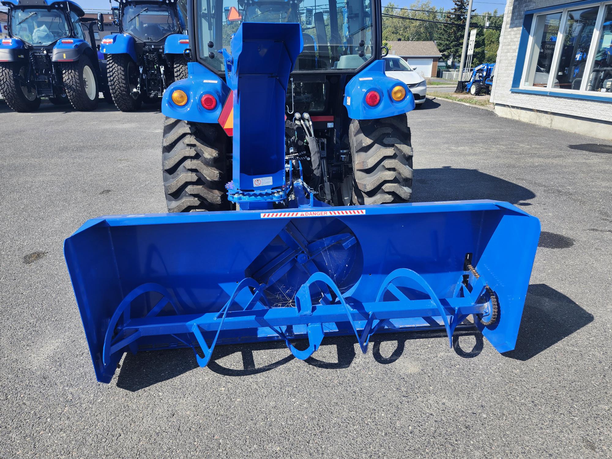 Une souffleuse à neige bleue est montée à l'arrière d'un tracteur, stationné sur un terrain pavé. Plusieurs autres tracteurs bleus sont visibles en arrière-plan près d'un bâtiment avec des fenêtres.